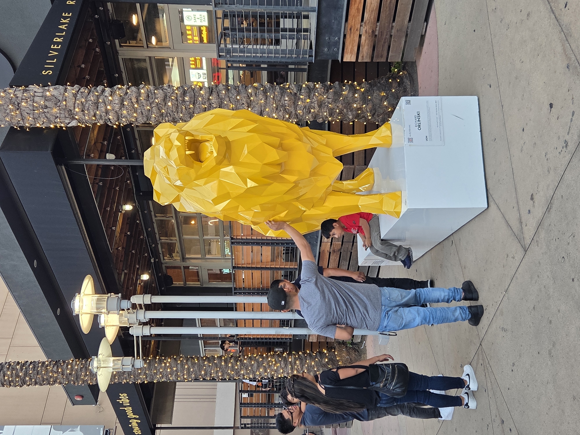 This is a full-shot, eye-level, outdoor photograph taken during the day on a paved sidewalk in front of a building with a black awning. To the left, there is a palm tree wrapped in small white lights. A yellow, faceted lion sculpture is the primary subject, standing on a white rectangular base. The sculpture is positioned to the right of the frame, with its head turned towards the viewer and its mouth open, revealing teeth. The building facade behind the sculpture has a sign that reads "SILVERLAKE R" in gold lettering. Several people are present in the foreground. On the left, a group of four individuals are standing, facing the sculpture. One person, wearing a dark shirt and jeans, is looking away from the camera. Next to them, a woman in a black top and jeans is looking at the sculpture. To their right, a man in a gray t-shirt and blue jeans is reaching out and touching the sculpture. Behind him, another person in a dark shirt is partially visible. To the right of the lion sculpture, a young boy in a red shirt and gray pants is sitting on the white base, looking at his phone. A small sign with text and QR codes is attached to the white base of the sculpture. The overall environment suggests an urban setting, possibly a public art installation or an outdoor seating area for a business.