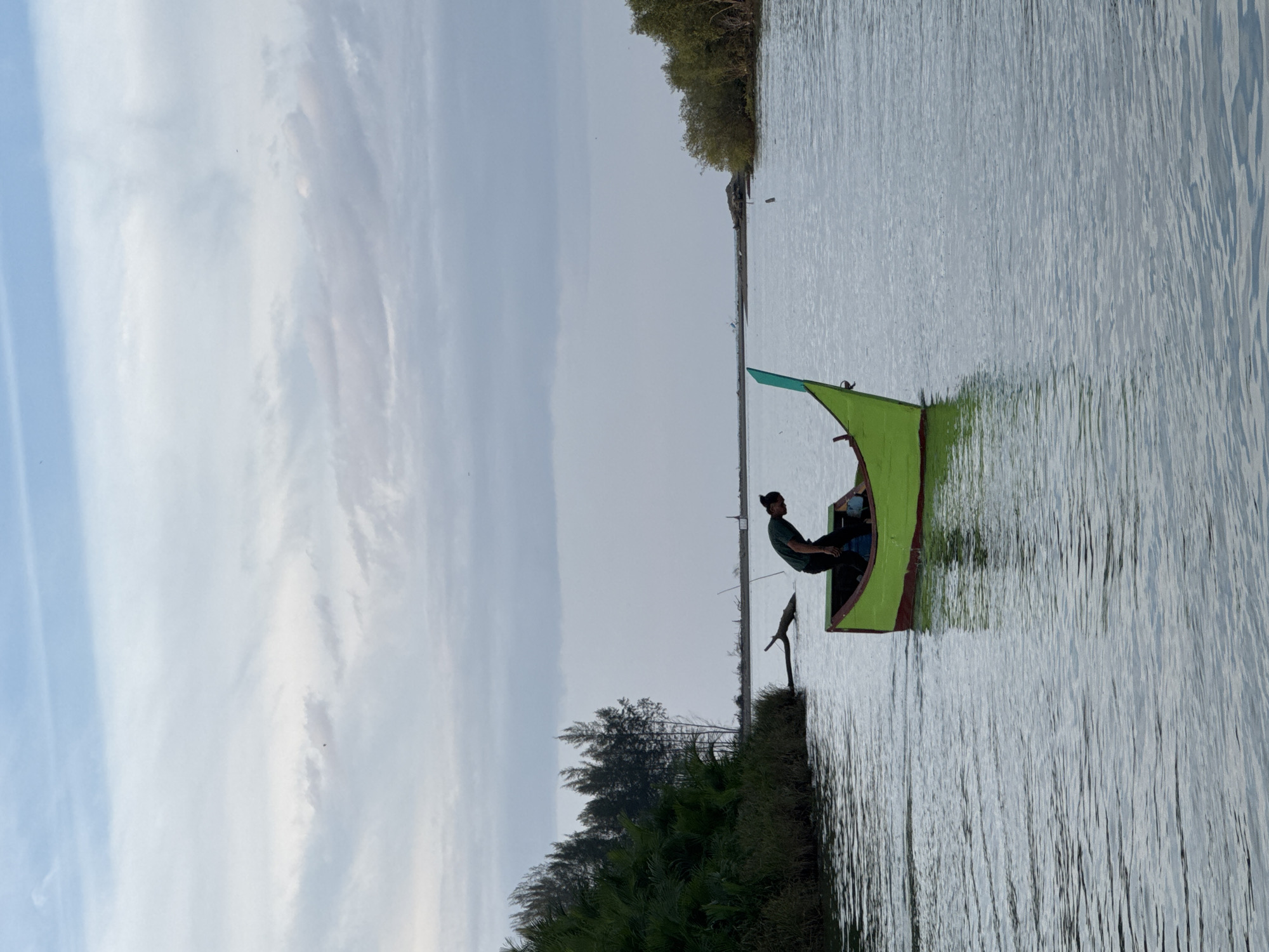 The image depicts a serene scene on a body of water, likely a river or bay, in Reudeuep, Indonesia. In the foreground, a person is bent over inside a small, brightly painted green and red boat, which has a distinctive pointed bow. The person is wearing a dark green shirt. The water is calm, with ripples indicating a gentle movement. To the left of the boat, the shore is lined with lush green vegetation and tall, slender trees. A piece of driftwood is visible in the water near the shore. In the background, the water stretches towards a distant horizon where the sky is filled with soft, grey and white clouds. The overall atmosphere is tranquil and natural.