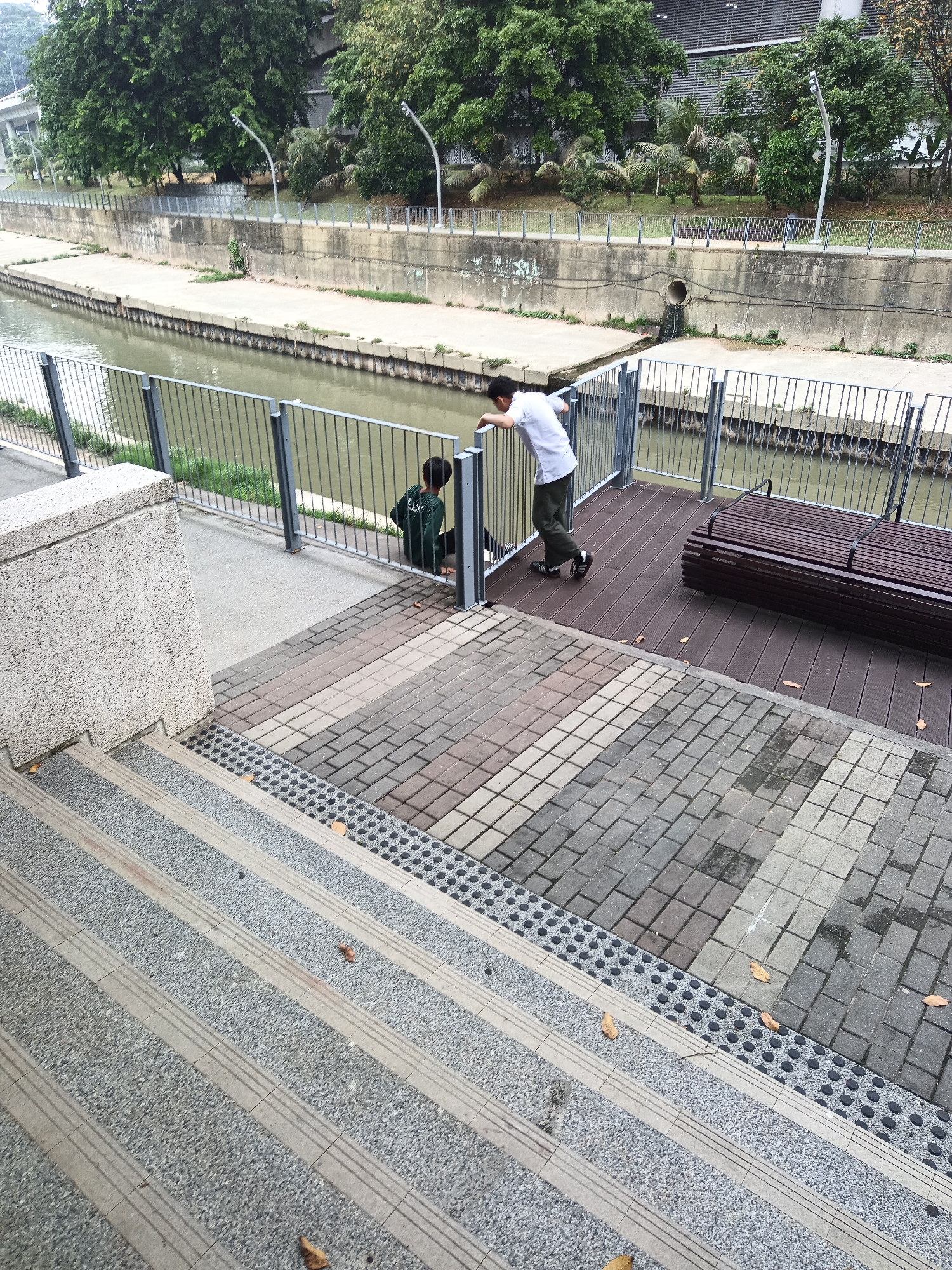 Two young men are by a canal in Kuala Lumpur, Malaysia. One man, wearing a white t-shirt and dark pants, leans over a metal railing. The other man, wearing a dark green long-sleeved shirt, sits with his back to the camera. The canal has a concrete wall and a metal fence along its edge. Adjacent to the railing is a wooden deck with a dark brown bench. In the foreground, there are wide concrete stairs with textured treads. Trees and buildings are visible in the background.