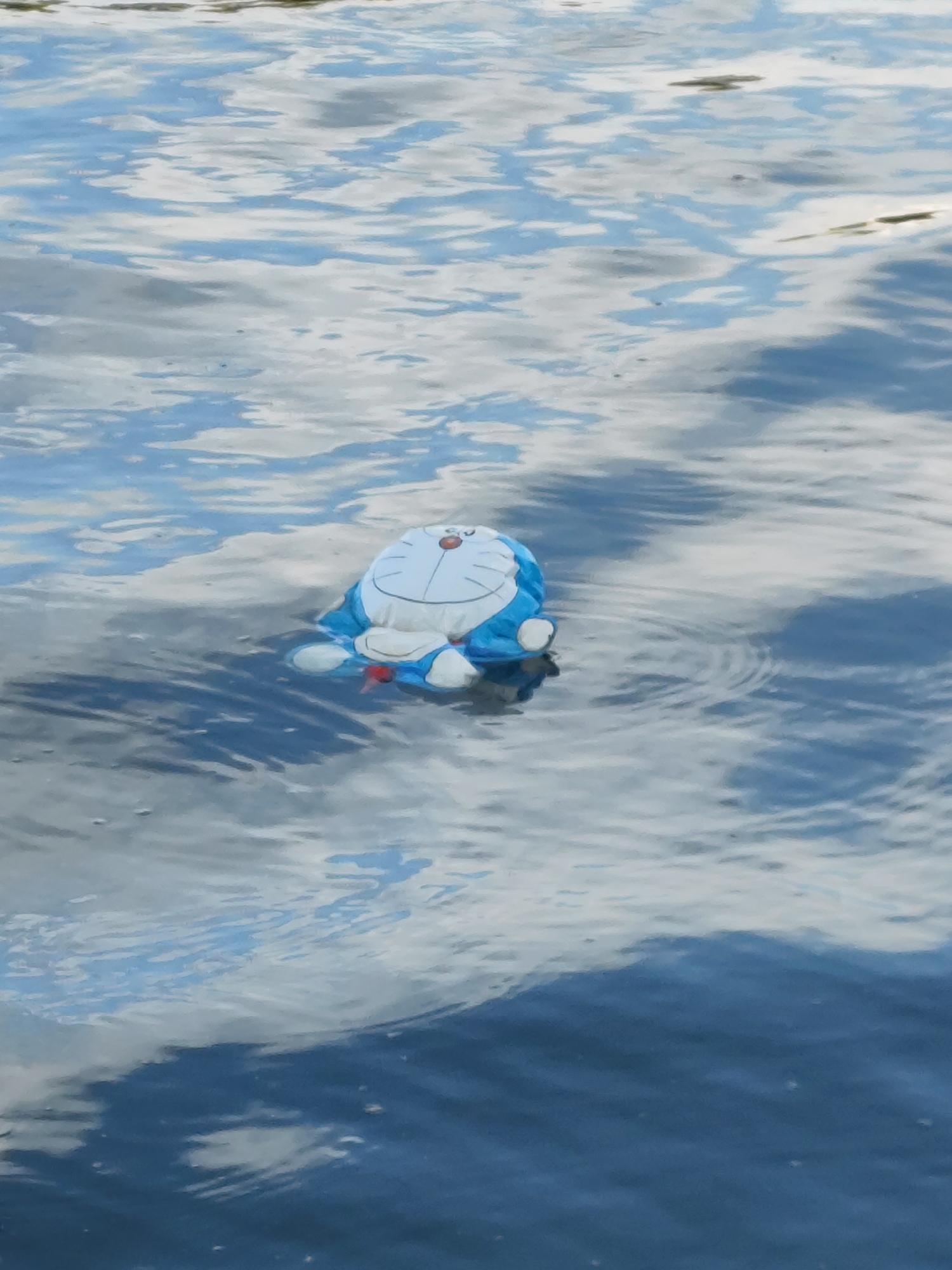 A stuffed Doraemon toy is floating on the surface of a body of water. The water has a rippled texture and reflects the sky, which appears cloudy with patches of blue. The Doraemon toy is primarily blue and white, with its signature red nose and a bell around its neck. It is positioned slightly off-center to the right. The surrounding environment is characterized by the water and the reflected sky. The scene is static and does not depict any people or specific actions beyond the toy floating. The location in Xuan Huong Lake, Da Lat, Vietnam, suggests an outdoor setting.