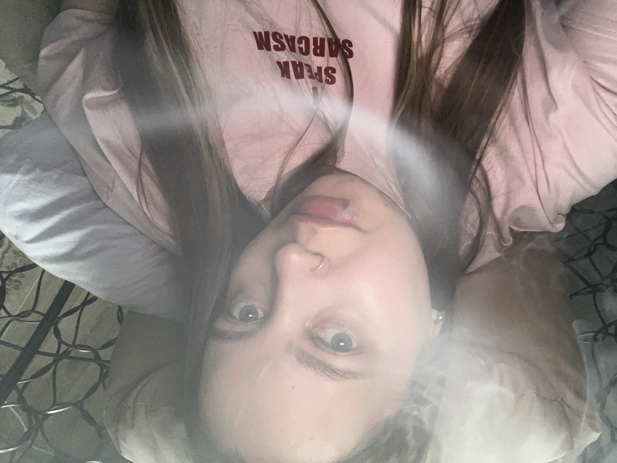 The image is a close-up, upside-down selfie of a young woman lying down. She has long, dark brown hair that falls around her face. She is wearing a light pink t-shirt with dark red text that reads "SPEAK SARCASM". The woman has wide, surprised-looking eyes and a small silver hoop in her nose. Her lips are slightly parted, and she appears to be looking directly at the camera. The background is soft and blurred, with hints of fabric and what appears to be netting or a string of lights. The overall impression is casual and perhaps a bit playful, given the t-shirt's slogan. The lighting suggests an indoor setting, possibly a bedroom, during the daytime. There are no specific indicators of the weather or a precise time of day, but the mood seems relaxed and intimate.