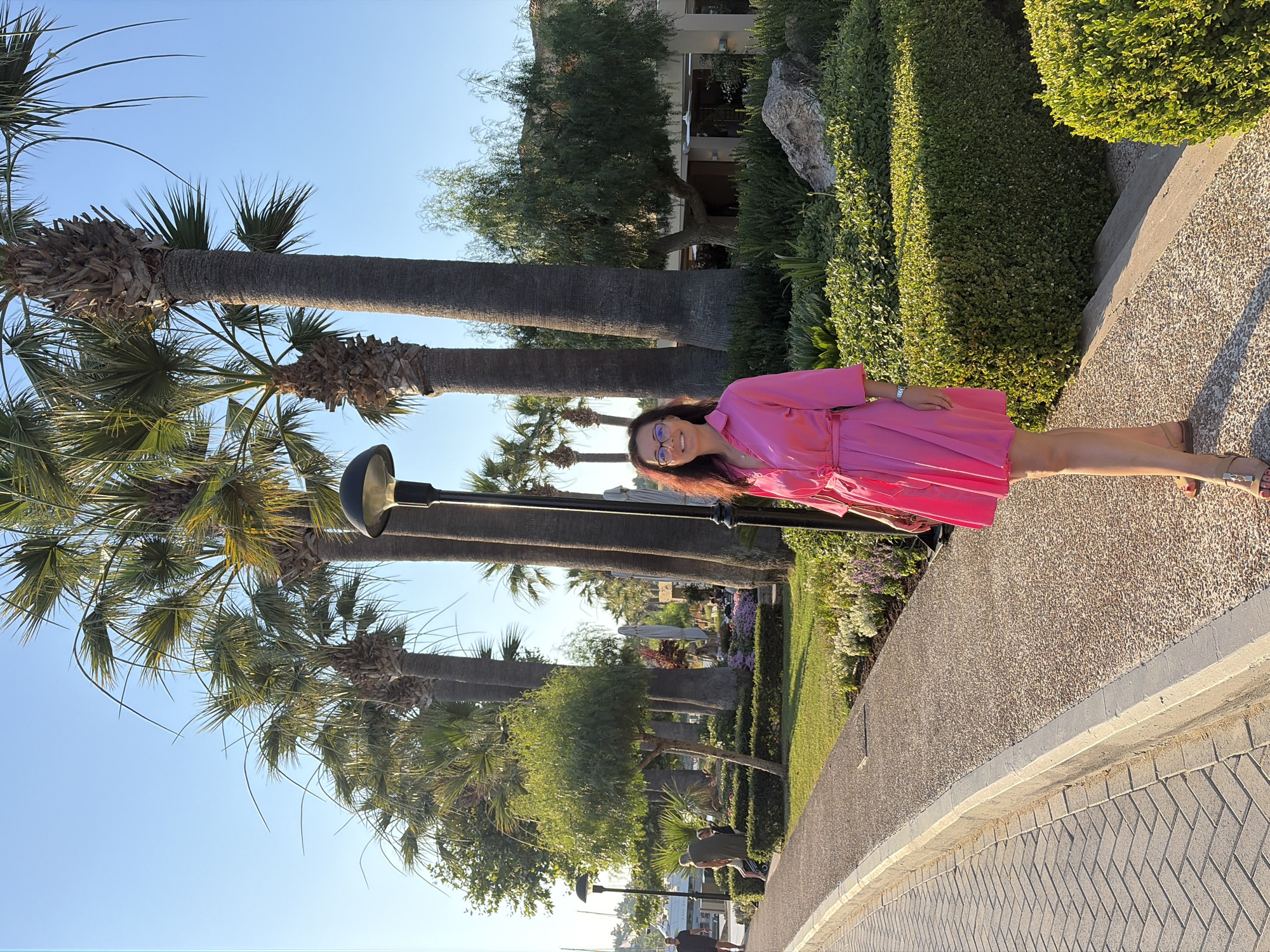 A woman in a bright pink dress and beige sandals stands on a paved walkway lined with manicured bushes and palm trees under a clear blue sky. She is smiling and appears to be enjoying a sunny day, likely in the late afternoon due to the warm lighting. The background features more lush greenery, some buildings with balconies, and a large rock. The walkway itself has a textured gravel surface next to a section of herringbone-patterned bricks. The scene suggests a warm, pleasant environment, possibly a resort or a well-maintained public space in a Mediterranean climate, consistent with the specified location of Kassandra Municipal Unit, Greece.