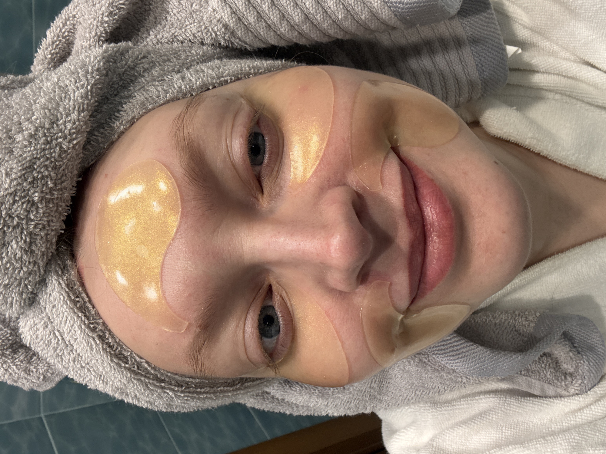 A woman lies on her back, wearing a gray towel wrapped around her head and a white towel around her neck. She has facial patches applied to her forehead, below her eyes, and around her mouth. Her eyes are open, and she is looking at the camera. There is no evidence to suggest the location is Izhevsk, Russia.