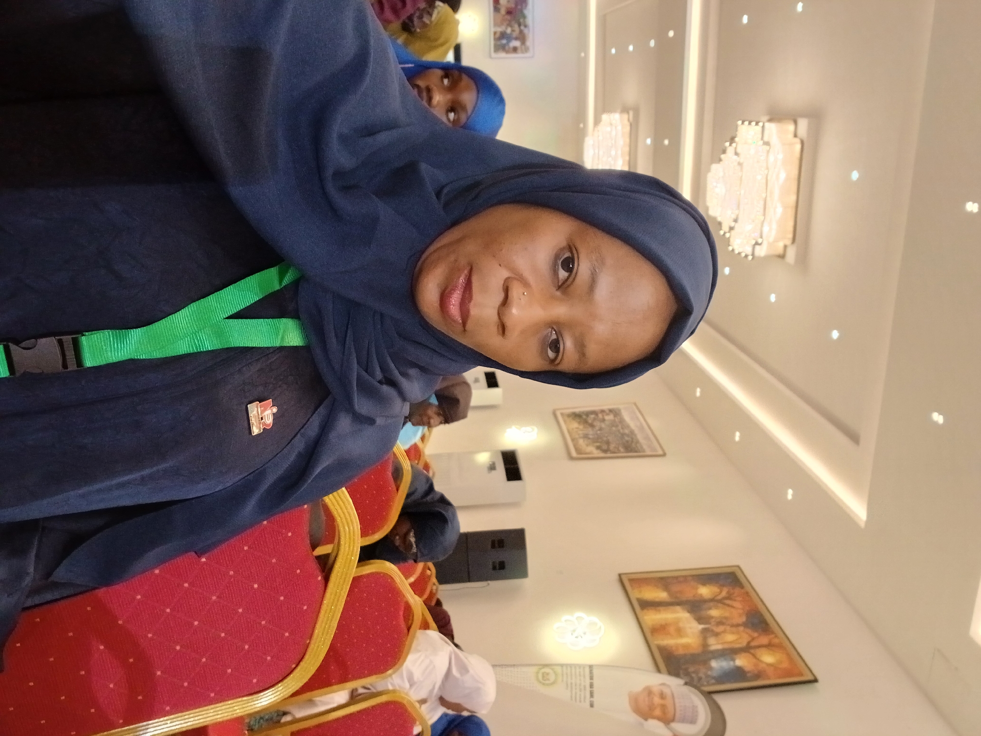 The image captures an indoor scene, likely a conference or event in Unguwan Sanusi, Nigeria. The primary subject is a woman in the foreground, wearing a dark blue hijab and a matching top, looking directly at the camera with a neutral expression. She has a nose piercing, and a bright green lanyard with a black plastic buckle is visible around her neck. A small red and white lapel pin is attached to her attire.
Behind her, another person in a blue hijab is partially visible, along with several other individuals seated in the background. The setting features rows of distinctive red chairs with golden frames. The room's decor includes light-colored walls and ceiling, adorned with decorative, ornate light fixtures resembling modern chandeliers, and framed artwork. White air conditioning units are mounted on the walls. In the far background, a large circular standee with a man's face and some unreadable text is visible. The overall impression is a formal gathering in a well-lit, contemporary event space.