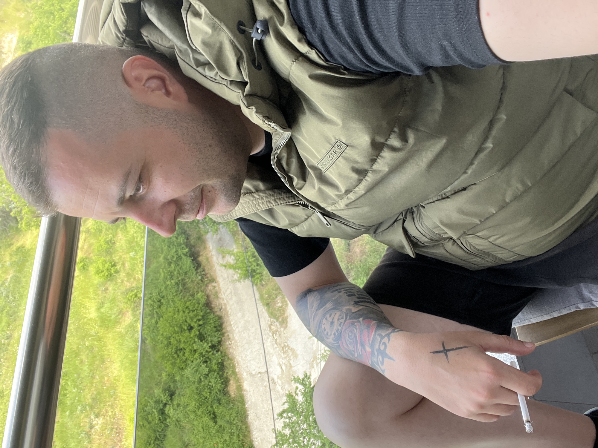 A man with short hair, wearing a green vest over a black shirt, is seated outside in Varna, Bulgaria. He is holding a lit cigarette in his right hand, which has a black cross tattoo. His left arm has a tattoo with a clock and a rose. A metal railing and green vegetation are visible in the background.