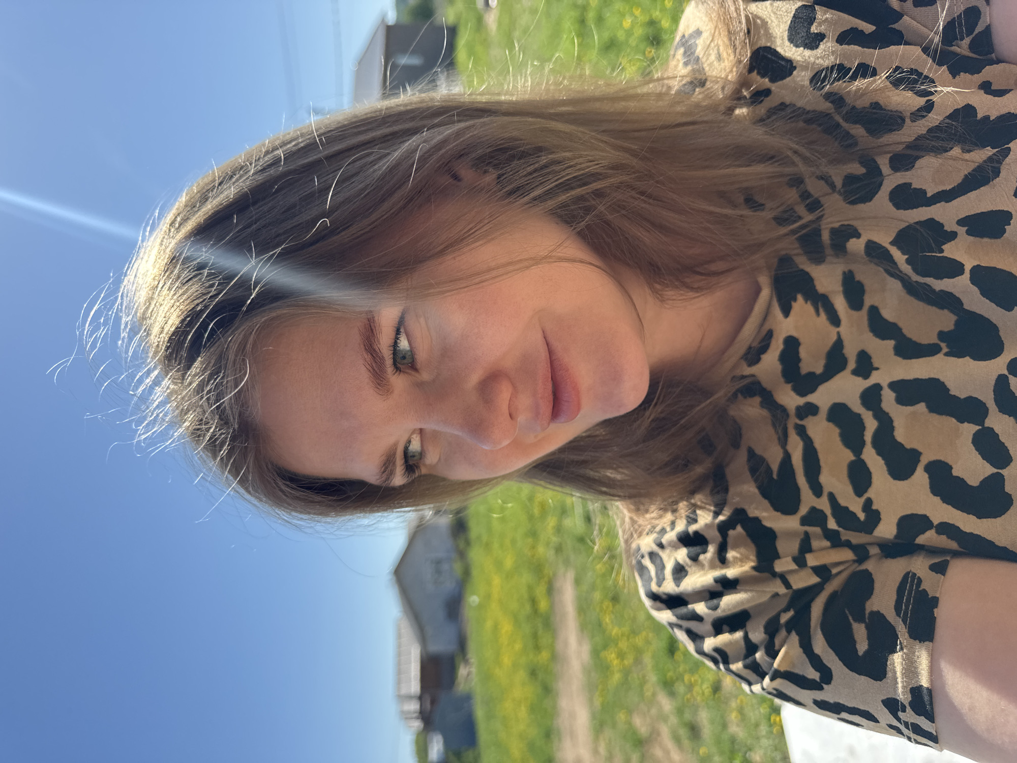 A medium, close-up, and slightly angled shot depicts a young woman with long, light brown hair and fair skin outdoors. She is wearing a brown and black animal print shirt. The background is bright, consisting of a clear blue sky and green grass with yellow wildflowers. A small, light-colored house is visible in the distance. A lens flare streak is present near the top left of the frame.
The location is Ферма, Russia.