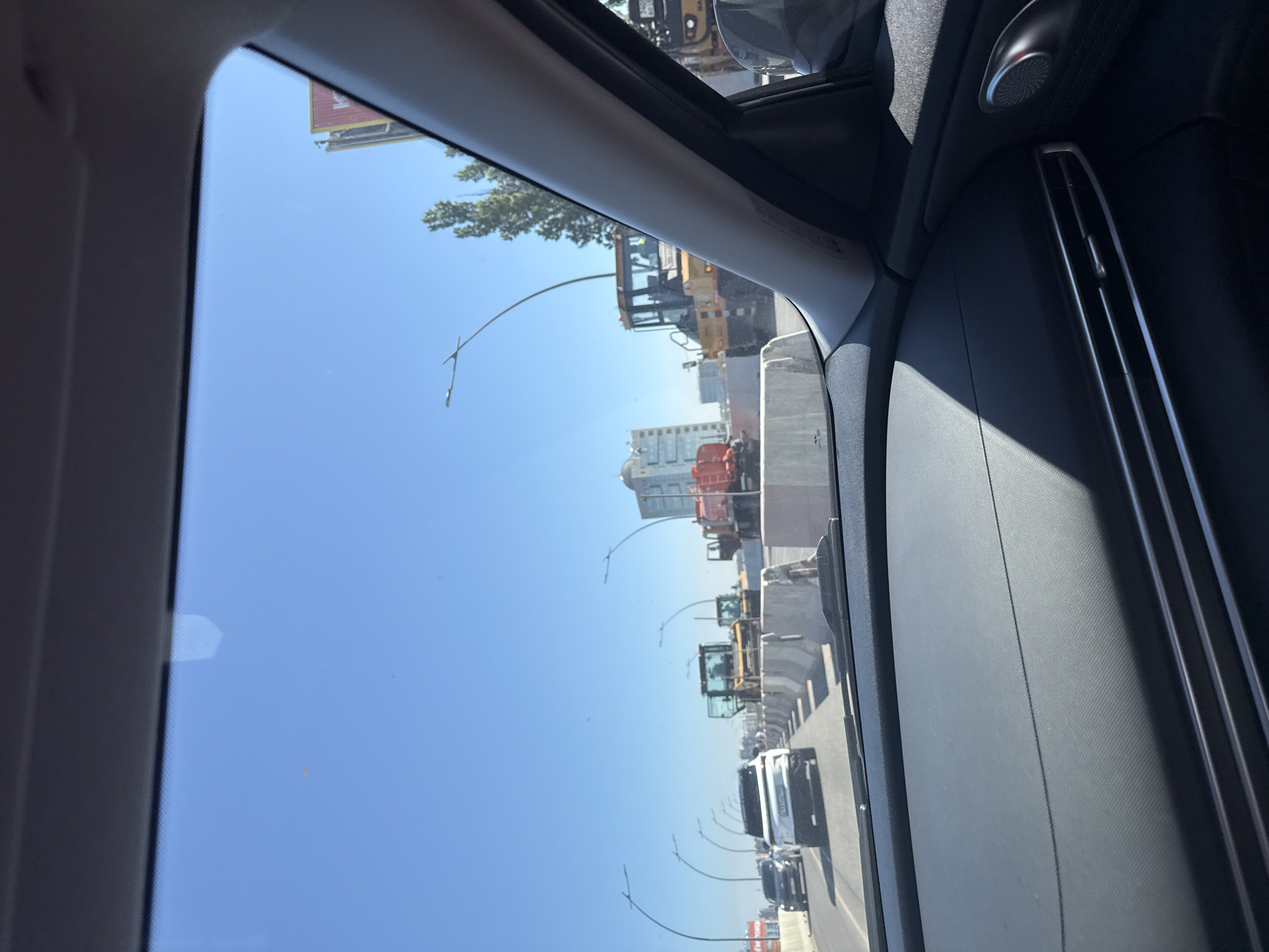 The image, taken from inside a car, shows an urban landscape on a clear, sunny day. The view is through the car window, framed by the car's interior, including the A-pillar and part of the dashboard. Outside the window, a busy road is visible, lined with streetlights, vehicles, and construction equipment. Several bulldozers and other heavy machinery are parked along the road. A high-rise building can be seen in the background.