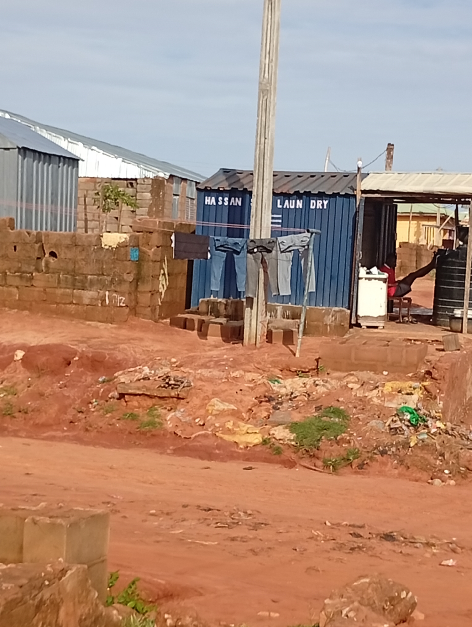 In Tumfure, Nigeria, a scene of daily life unfolds against a backdrop of utilitarian structures and weathered earth. A blue shipping container, repurposed as a laundry business, stands prominently with "HASSAN LAUNDRY" emblazoned on its side. Clothes hang drying on a line strung in front of it, their colors muted by the harsh sunlight. A white washing machine is visible through an open doorway. Inside the laundry, a person in a red shirt and dark shorts sits on a stool, facing away from the camera, appearing to be taking a break or tending to operations. Adjacent to the laundry, a large black water tank is visible. The ground is a dusty, reddish-brown earth, scattered with debris, indicating a place of industry and commerce in a possibly developing area. The surrounding buildings are constructed from corrugated metal and rough-hewn bricks, reflecting a pragmatic approach to construction. A solitary wooden pole, likely supporting utility lines, stands tall against a pale blue sky. The overall impression is one of a functioning, though perhaps modest, business operating in a rural or semi-urban Nigerian setting.