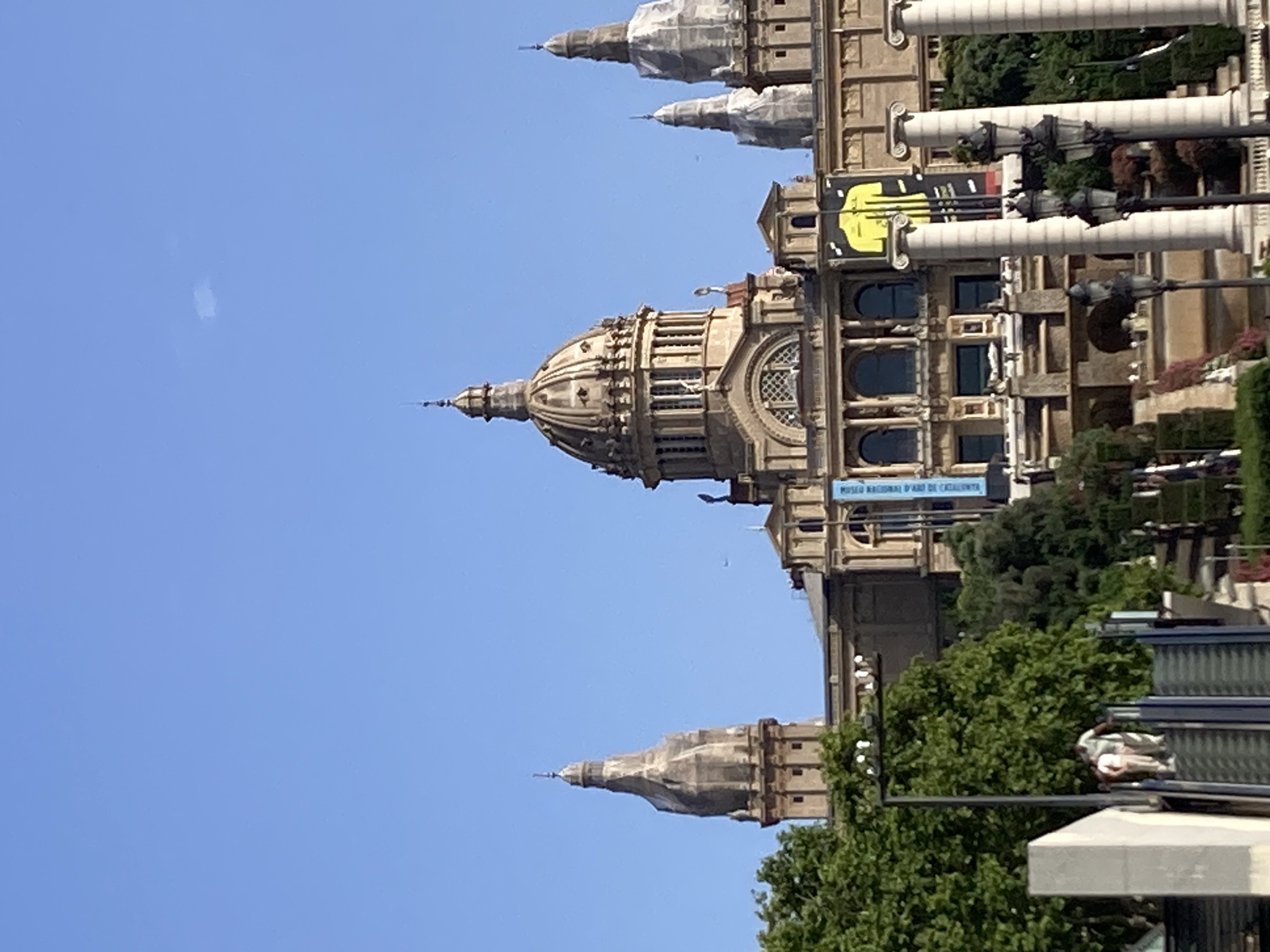 The image shows the Palau Nacional, a monumental palace located on Montjuïc hill in Barcelona, Spain. The palace is the home of the Museu Nacional d'Art de Catalunya (MNAC). The building features a prominent central dome and is flanked by two towers, with additional smaller domes and spires visible. A wide, ornate staircase leads up to the palace, with statues and fountains adorning its sides. Lush green trees are visible in the foreground and to the sides of the palace. The sky is a clear, bright blue. In the lower left portion of the image, two people are visible on an escalator. There are also decorative lampposts and columns in the foreground. Two large banners, one blue and one yellow, are hanging on the facade of the palace.
