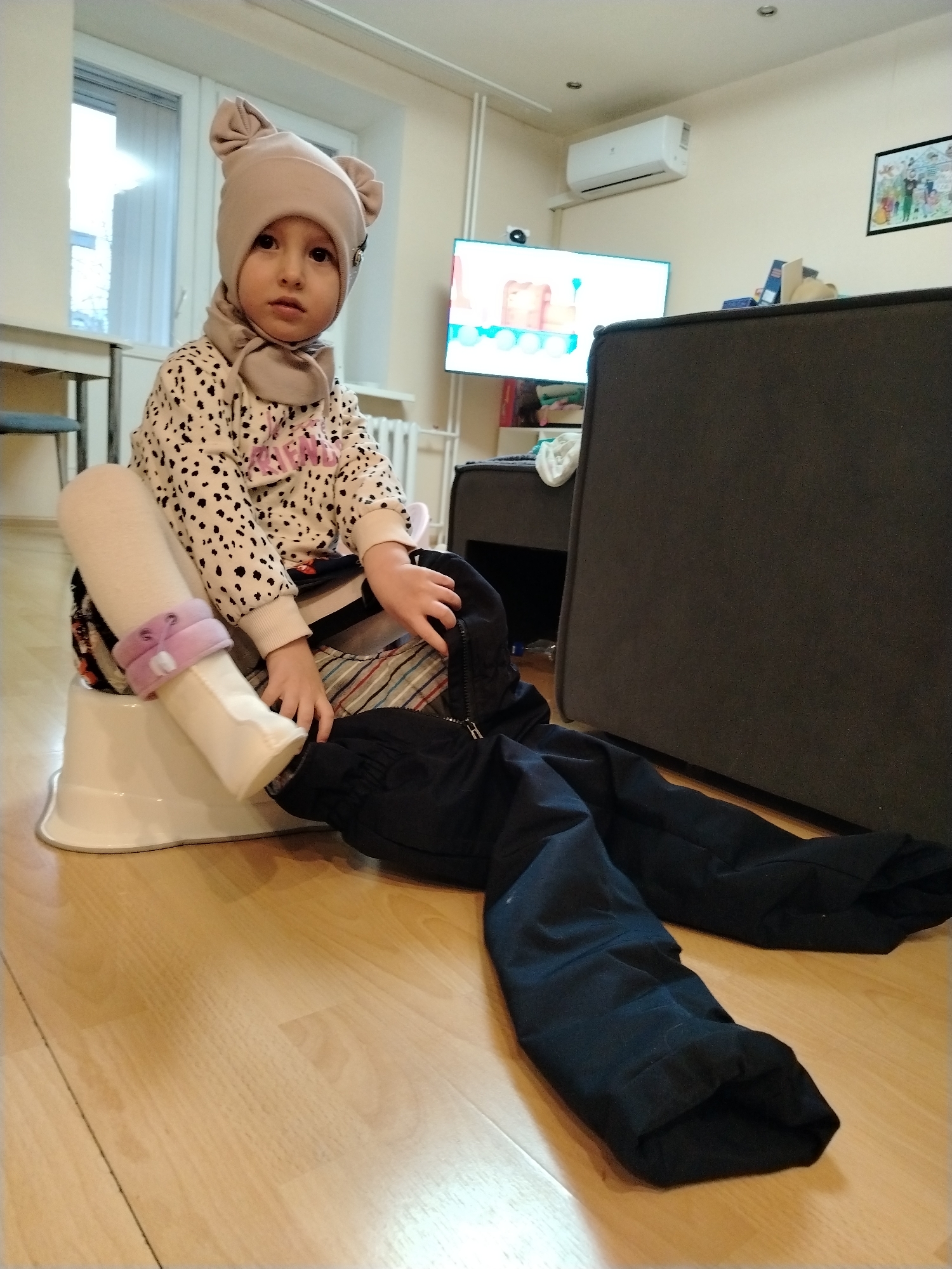 A young child wearing a beige head covering with attached scarf, and a long-sleeved shirt with black spots and pink writing, is sitting on a white potty chair on a light-colored wooden floor. The child's left leg, clad in a white stocking with a pink decorative band, is extended forward. Beside the potty, a pair of dark-colored snow pants lay on the floor.
In the background, a television is visible on a white shelving unit. Behind that is a grey couch. A window with blinds is located on the left, and an air conditioner is visible on the wall toward the top. A framed picture is on the wall.
The picture was likely taken indoors, in an apartment located in the city of Chaikovsky, Russia.