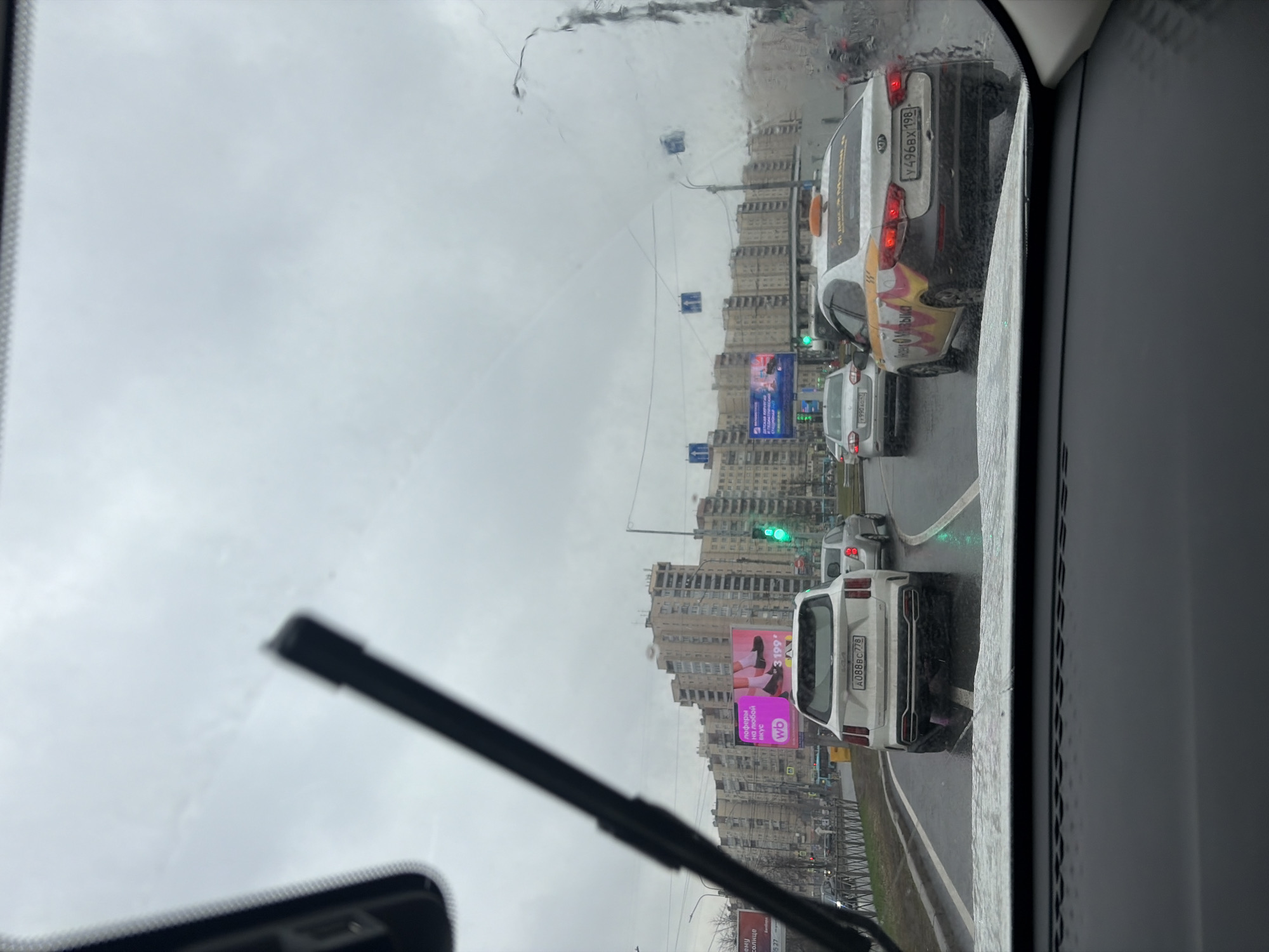 A view from inside a car, looking through a rain-splattered windshield, shows a city street scene. Cars are in traffic, and the buildings lining the street are tall and residential-looking. There are advertisements on the side of the road, as well as traffic lights. The image seems to have been taken in Saint Petersburg, Russia, as the license plates on the cars have the Russian Federation flag on them.