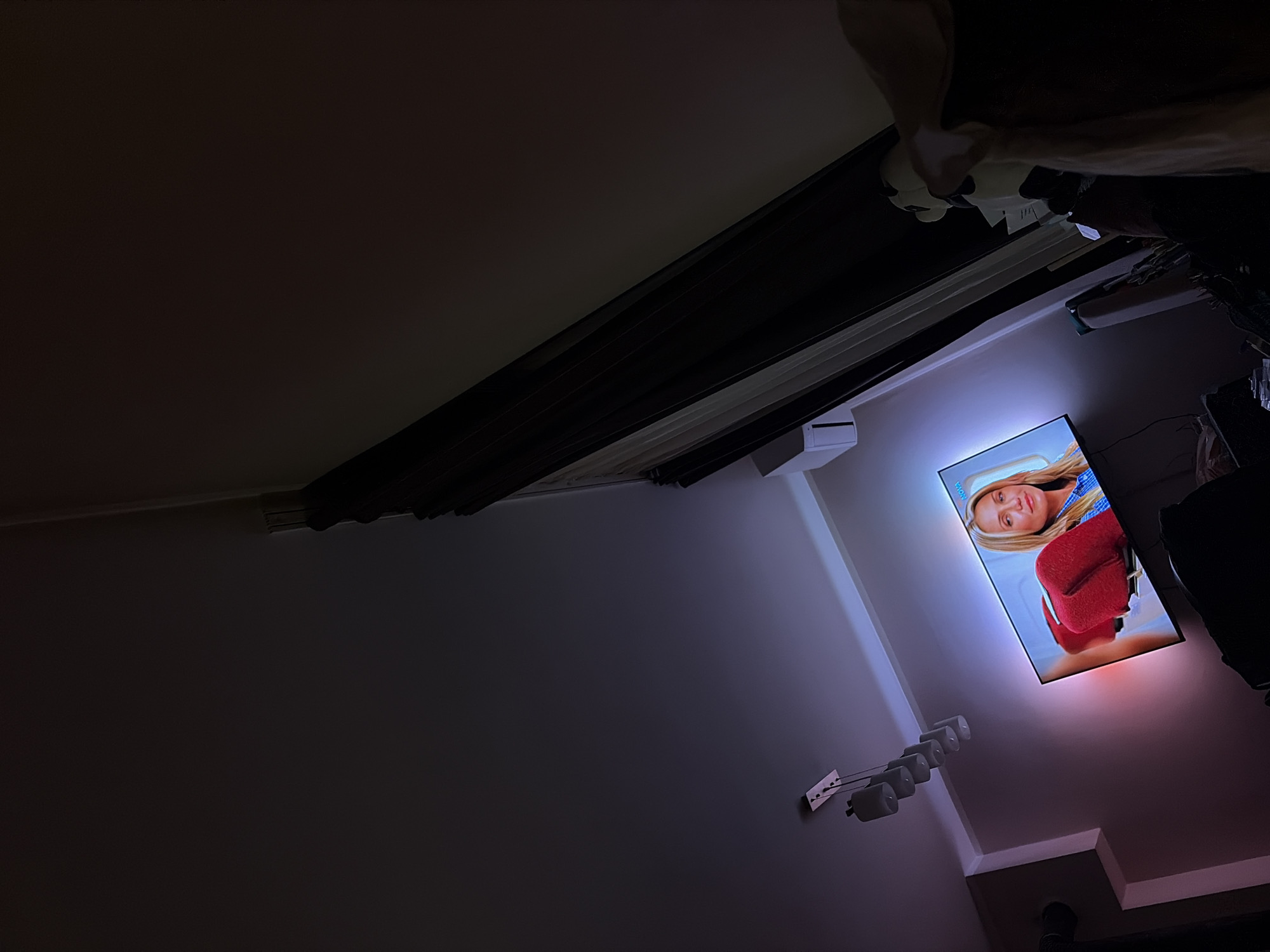 This image depicts an interior residential space in Tsarevo, Bulgaria, under dim lighting conditions. A large, wall-mounted flat-screen television is centrally positioned, displaying a blonde woman with a neutral expression, wearing a blue and white striped top. A prominent red, padded object, resembling an aircraft seat component, is visible in the foreground of the screen. The television screen displays the "NOVA" logo in its upper right corner and emits ambient backlighting that casts purple and pink illumination onto the surrounding wall.
Above the television, a contemporary ceiling light fixture with five grey cylindrical lamps is suspended. To the upper left of the television, a white wall-mounted air conditioning unit is present. The walls are light-colored, exhibiting color variations due to the television's backlighting. Dark, floor-length curtains are drawn along the right side of the frame, covering a window or doorway. In the lower right corner, indistinct dark objects, possibly furniture, are partially visible, along with a light-colored textile and a white and black stuffed animal. A dark vertical pipe or conduit is visible in the upper left section of the room.