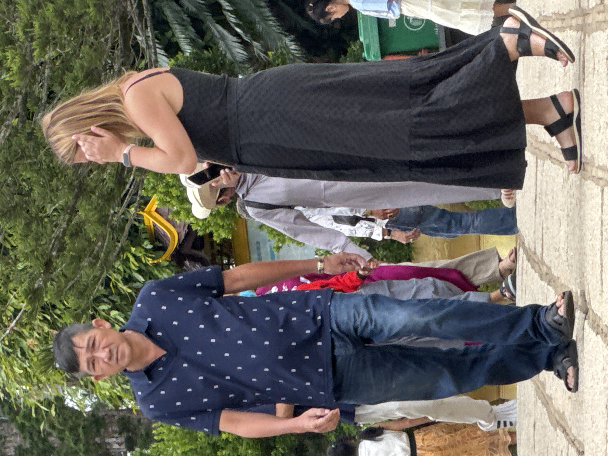 The image shows a group of people walking on a paved pathway lined with trees. In the foreground, a man in a dark blue polo shirt with a white polka dot pattern and blue jeans is walking towards the viewer. He is wearing black sandals. To his right, a woman in a long black dress and black sandals is walking away from the viewer, with her hand covering part of her face. Behind them, several other individuals are visible, some in traditional Vietnamese clothing. In the background, there are lush green trees and a glimpse of a yellow structure. The pathway appears to be in an outdoor setting, possibly a tourist attraction or a temple complex, consistent with locations in Da Lat, Vietnam.