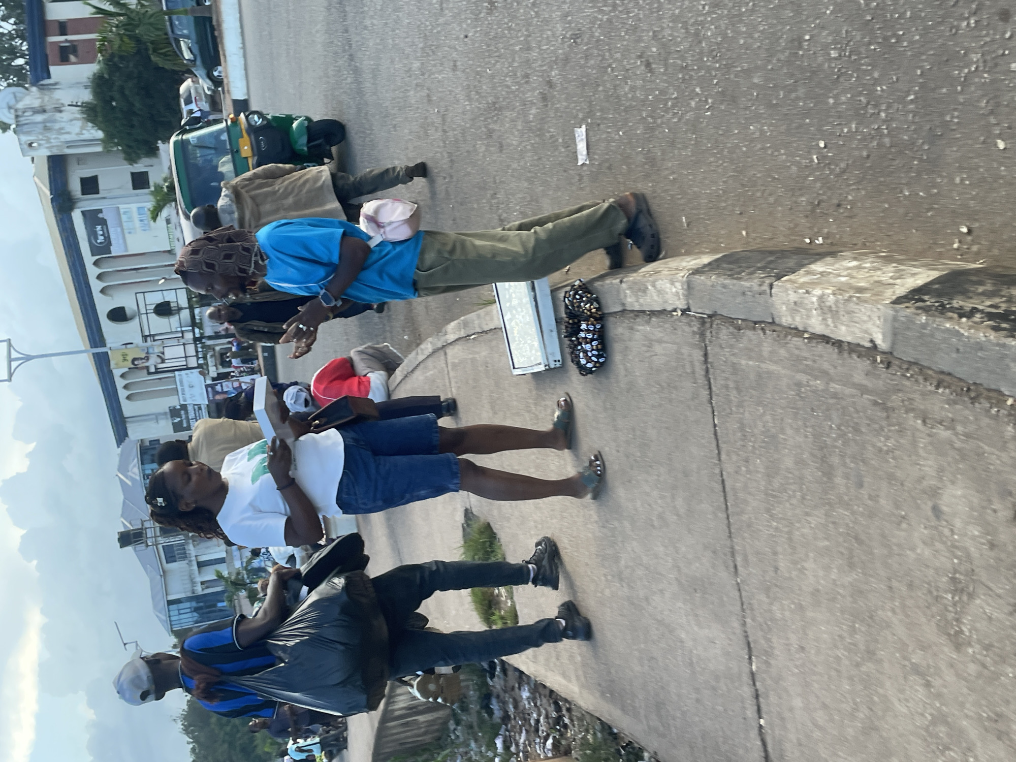 In the city of Jos, Nigeria, an outdoor scene unfolds on a sidewalk adjacent to a paved road. The time of day appears to be late afternoon, indicated by the soft, diffused lighting and the shadows cast by the subjects.
In the foreground, two individuals are engaged in an interaction. A woman, wearing a white t-shirt and blue shorts, stands on the sidewalk holding a white box. She appears to be presenting it to a person facing her. The person facing her, dressed in a blue t-shirt and olive green pants, stands on the edge of the sidewalk, facing the woman. They are looking at each other, and their interaction suggests a transaction or exchange. The person in blue has a light-colored bag slung over their shoulder. Beside the person in blue, on the curb, a flat, rectangular object and a patterned bundle of small items are placed, possibly merchandise for sale.
Behind the central figures, a man in a blue and black striped shirt with a white cap walks away from the camera, carrying a large black plastic bag over his left shoulder. Further in the background, other people are visible, some walking, some possibly sitting. A green motorized tricycle, often used as a taxi in Nigeria, is parked on the road, along with other vehicles. The background also shows buildings, some of which have architectural features that suggest they are commercial establishments or offices. Trees and overhead utility lines are also present in the environment. The overall impression is of a busy urban street with pedestrian and vehicular traffic.
