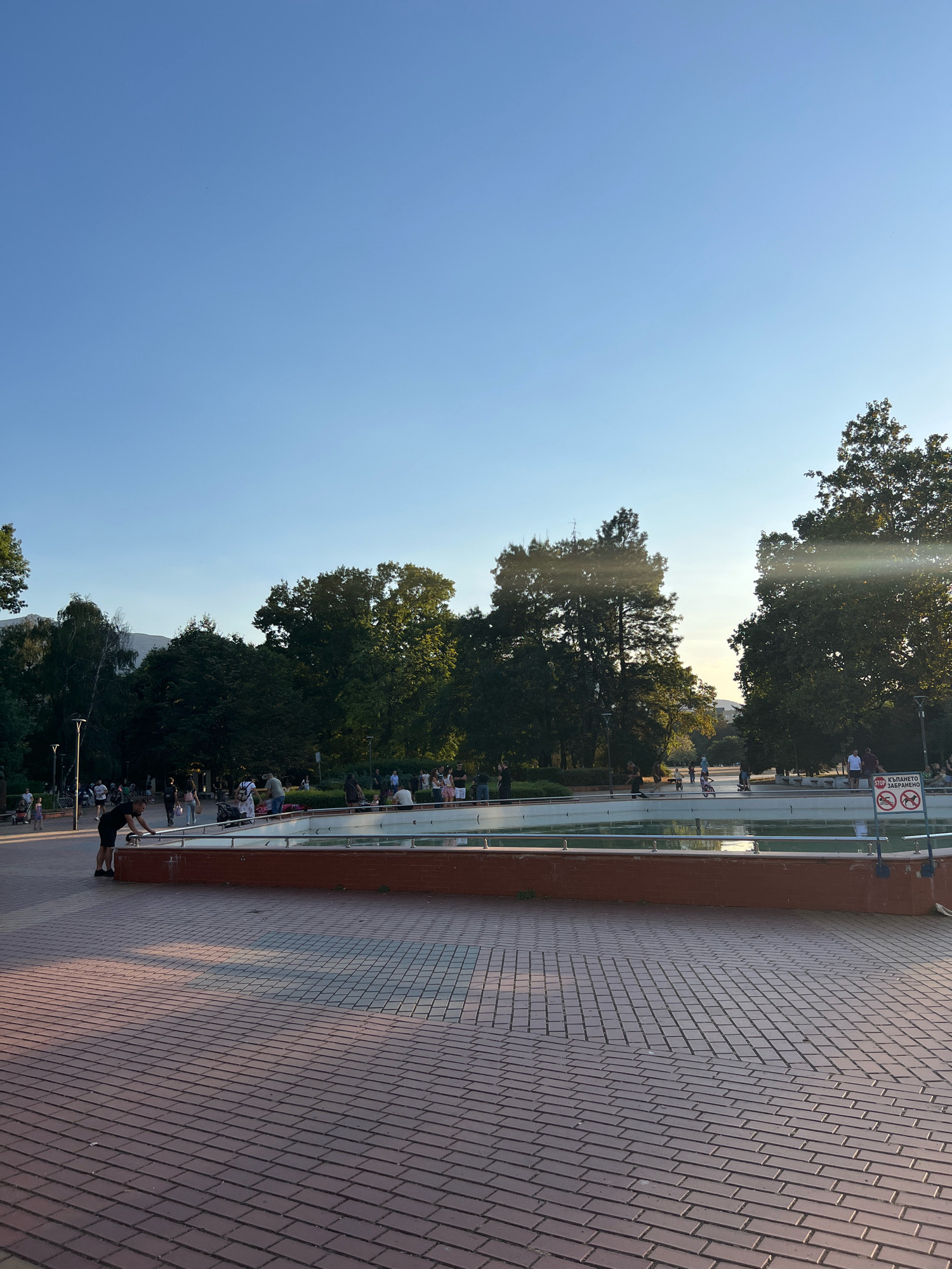 The image depicts a public park with a dry fountain in the foreground. The fountain's basin is a light blue color and is surrounded by a brick structure with a metal railing. Beyond the fountain, a paved pathway is visible, with many people walking and standing around. The pathway leads to a tree-lined area with more people. The trees are lush and green, suggesting it is summer or late spring. The sky is clear and blue. In the background, there is a hint of mountains. To the right of the fountain, there is a sign with Bulgarian text and a prohibition symbol that appears to depict dogs and a person swimming. The lighting suggests it is late afternoon or early evening, with the sun casting long shadows. The overall scene appears to be a lively public space. This scene is likely from the Borisova Gradina park in Sofia, Bulgaria, which features a prominent dry fountain and numerous recreational areas.