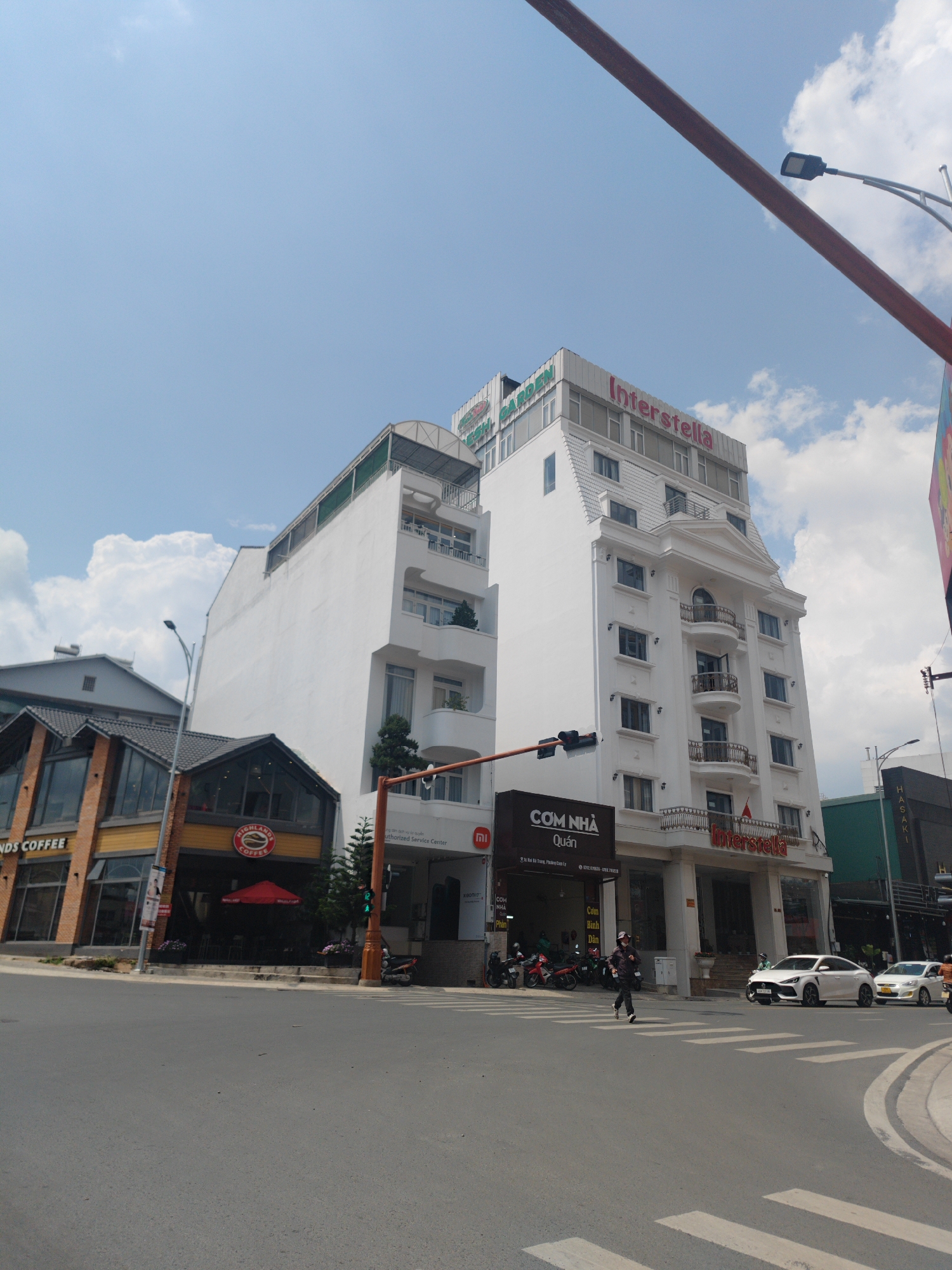 The image shows a street corner in Cam Ly - Đà Lạt, Vietnam. On the left is a tilted two-story building, housing "Highlands Coffee," recognizable by its brown and yellow color scheme. Next to it, there is a taller, rectangular white building with multiple stories. Above the entrance, there is a sign that reads, "Cơm Nhà Quán." The word "Interstella" is visible on the top of the building. A person is crossing the street in the crosswalk, and several parked motorcycles are nearby. The sky is partly cloudy. Two cars are present on the right side of the image. A red support beam cuts diagonally through the upper right quadrant of the photo.