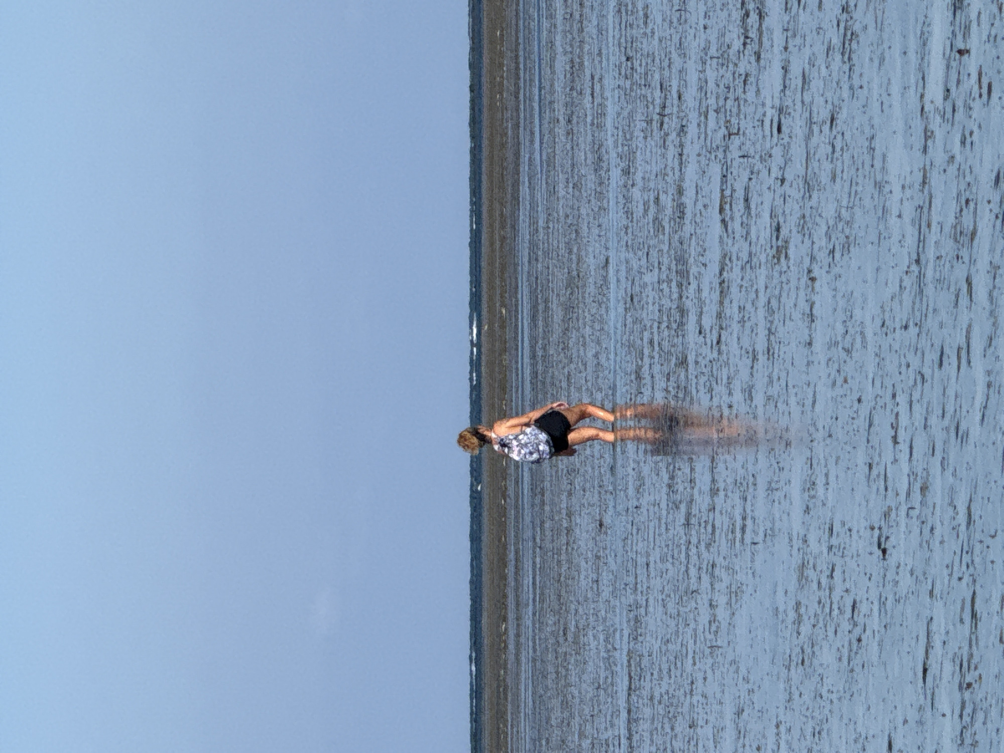 A person with blonde hair, wearing a black and white patterned backpack and black shorts, stands in shallow water on a muddy, flat expanse. The person is facing away from the camera, with their body angled slightly to the right. The water reflects the blue sky and the person's legs. In the distance, a dark line of the sea is visible, with a thin strip of pale sand or land beyond it. The sky is a clear, pale blue with a few faint clouds near the horizon. The overall impression is one of a vast, open, and serene environment.