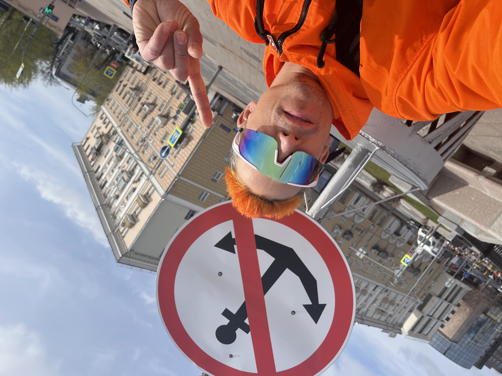 A person wearing an orange jacket and futuristic-looking sunglasses, with orange hair, is pointing at a round, white-and-red traffic sign depicting an anchor crossed out by a red line. The sign is presumably indicating that anchoring is prohibited. The person appears to be outside, in what looks like a city environment. The background shows buildings and street signs. The building's architecture and signage suggest that the location is Moscow, Russia.