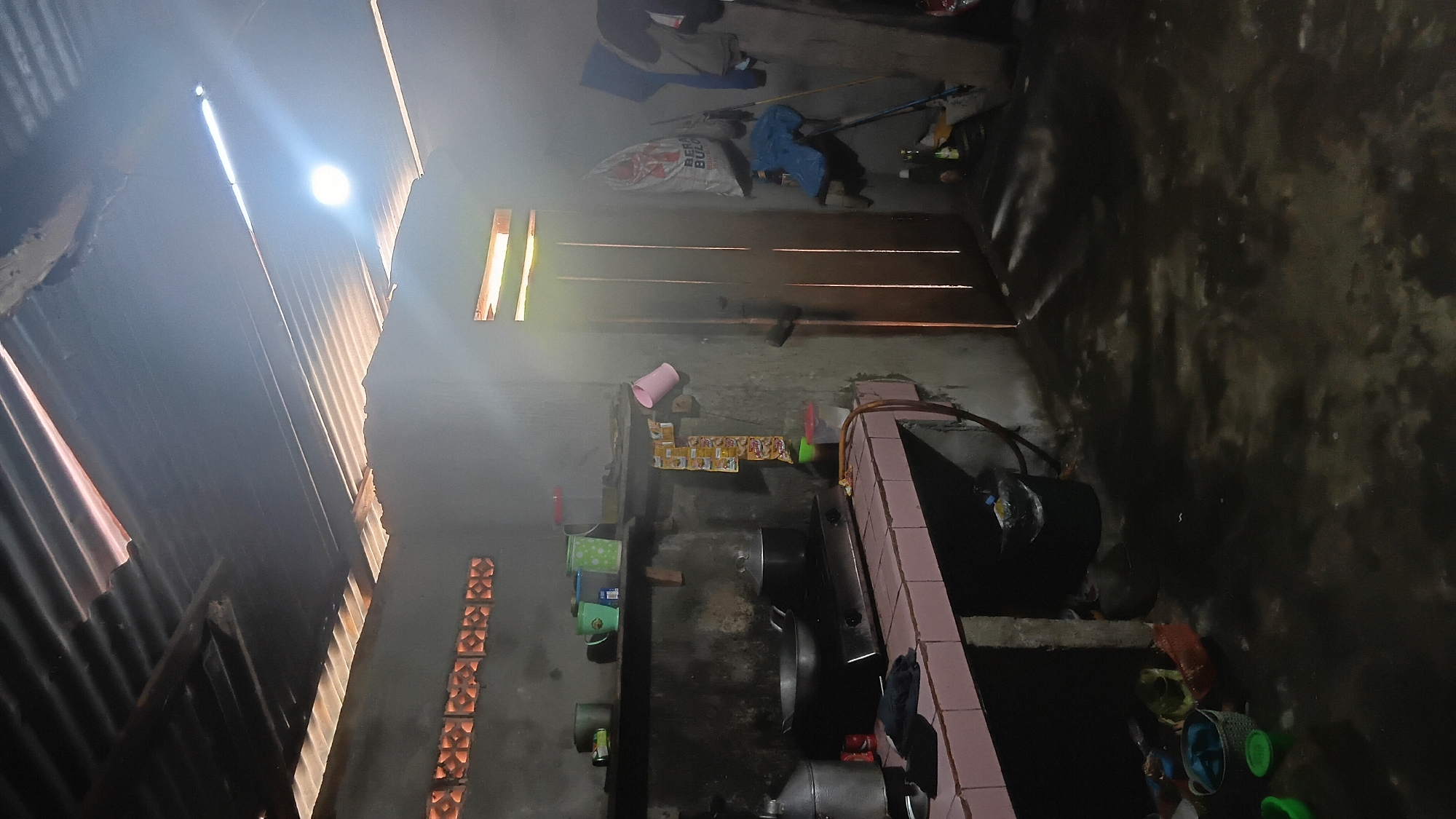 The image depicts an interior view of a rustic kitchen, likely in Prabumulih, Indonesia, characterized by its simple construction and minimal amenities. The ceiling is made of corrugated metal, with visible gaps and some light filtering through, creating a diffused lighting effect. The walls appear to be rough concrete, with some sections showing exposed brickwork or ventilation blocks.
In the foreground, a tiled countertop with a pink trim serves as the primary workspace. On it, a gas stove with two burners is present, with a cooking pot and a lid on one of the burners. To the left of the stove, a large metal container, possibly for water or storage, is visible. Several smaller containers, cups, and what appear to be sachets of seasoning or instant noodles are arranged on a shelf above the countertop. A pink mug hangs from the shelf.
To the right of the kitchen area, a wooden door with vertical planks is centrally located, allowing natural light to stream in, creating streaks of light on the floor and walls. The door is slightly ajar. To the right of the door, several items are hanging, including a white sack with text that is partially obscured, a blue bag, and some articles of clothing. A broom and a mop lean against the wall near the door. On the floor near the door, there are several bottles and containers.
The floor is a dark, wet surface, possibly concrete, with visible puddles and reflections of light. To the left of the countertop, a black trash bin is situated. Below the countertop, a collection of various containers, including baskets and plastic basins, are placed on the floor.
The overall atmosphere suggests a functional, lived-in kitchen, possibly an outdoor or semi-outdoor kitchen, common in some residential areas. The presence of cooking utensils, storage items, and cleaning tools indicates daily use. The lighting suggests it is either early morning or late afternoon, with the sun's rays penetrating the space. There are no people visible in the image.