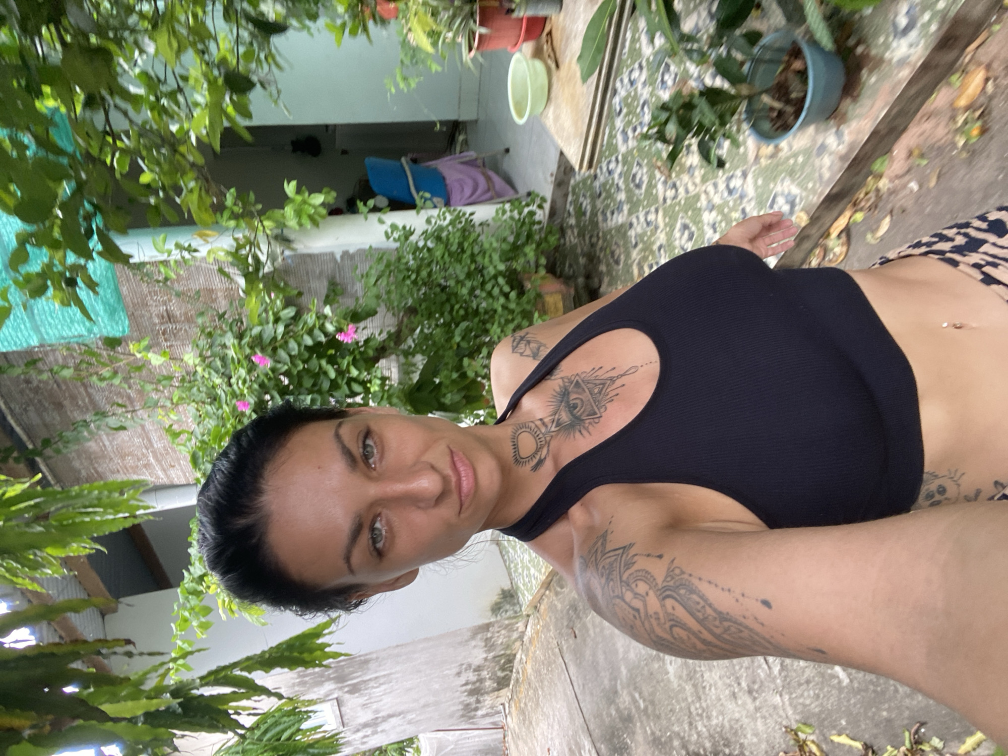 A woman is taking a selfie in a courtyard. She has dark hair pulled back and green eyes. She is wearing a black sports bra and patterned shorts. She has tattoos on her arm and chest. The courtyard is paved with concrete and decorated with potted plants. There is a building in the background. The overall setting suggests a residential or outdoor space in Mũi Né, Vietnam.
