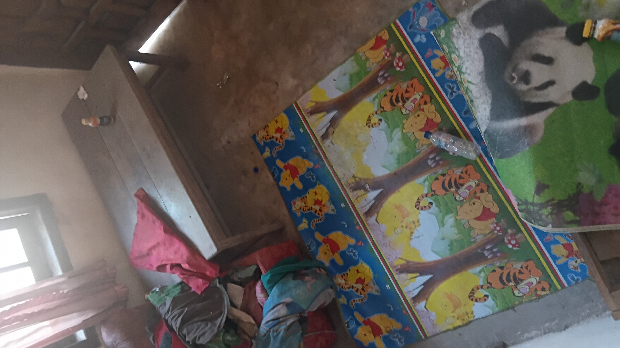 The image depicts a cluttered indoor scene, possibly a bedroom or living area, with objects scattered across the floor and a wooden table. In the foreground, a colorful children's mat featuring Winnie the Pooh and Tigger characters is laid out. Next to it, another mat with a panda design is partially visible. A plastic water bottle lies on the Winnie the Pooh mat. A wooden table occupies the mid-ground, with a red cloth draped over it and a small bottle with a yellow cap placed on its surface. To the left of the table, a pile of assorted clothing and fabric items is visible, including a red piece of cloth. The background shows a window with pink curtains and a wooden door. The floor appears to be concrete. The scene suggests a lived-in space, possibly in a residential area in Prabumulih, Indonesia, given the context provided. There are no individuals present in the image.