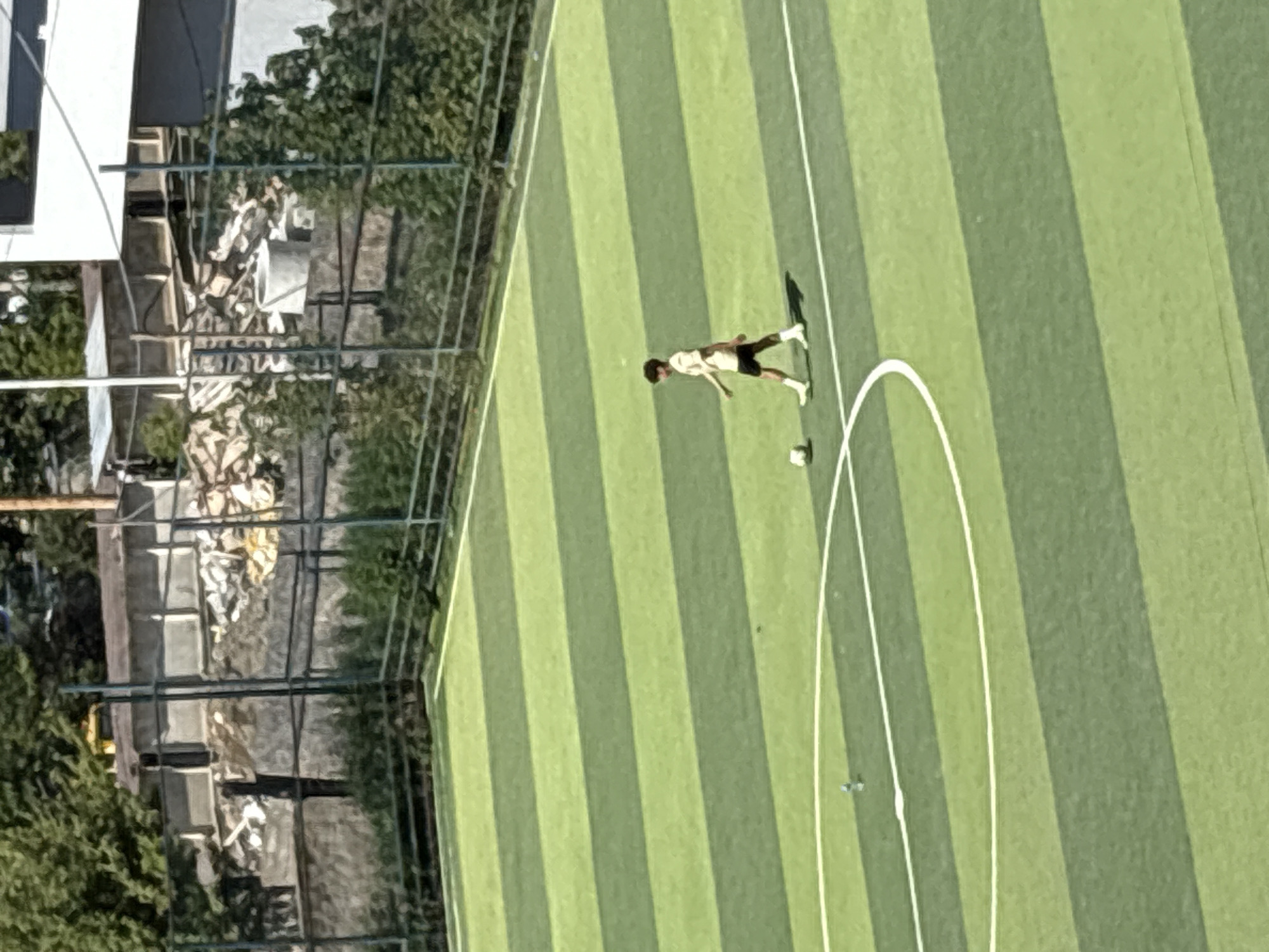 A lone young male, appearing to be a teenager, is walking across a green and lime-striped artificial turf soccer field. He is wearing a light-colored t-shirt, dark shorts, white socks, and white athletic shoes. A soccer ball is positioned to his left, approximately two feet in front of him. The field is bordered by a dark green chain-link fence. In the background, beyond the fence, there are trees and buildings under construction. The scene appears to be in a city. The presence of construction and the general urban environment suggests this could be an area undergoing development in Tbilisi, Georgia.