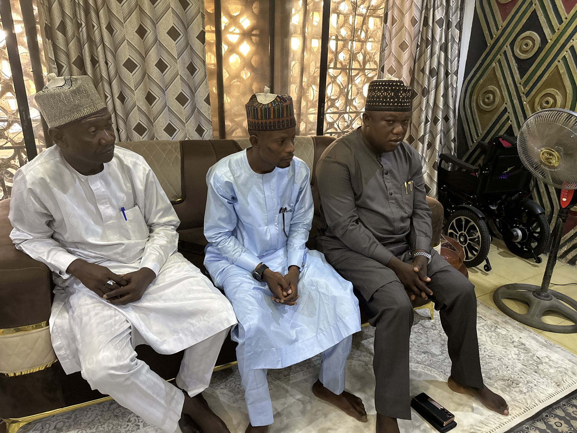 Three men are seated on a brown sofa. The man on the left wears a white outfit and a light-colored cap. The man in the middle is in light blue clothing with a patterned cap. The man on the right wears a grey suit and a dark cap. All three are barefoot. Behind them are patterned curtains, a wheelchair, and a standing fan. The location is Unknown, Unknown.