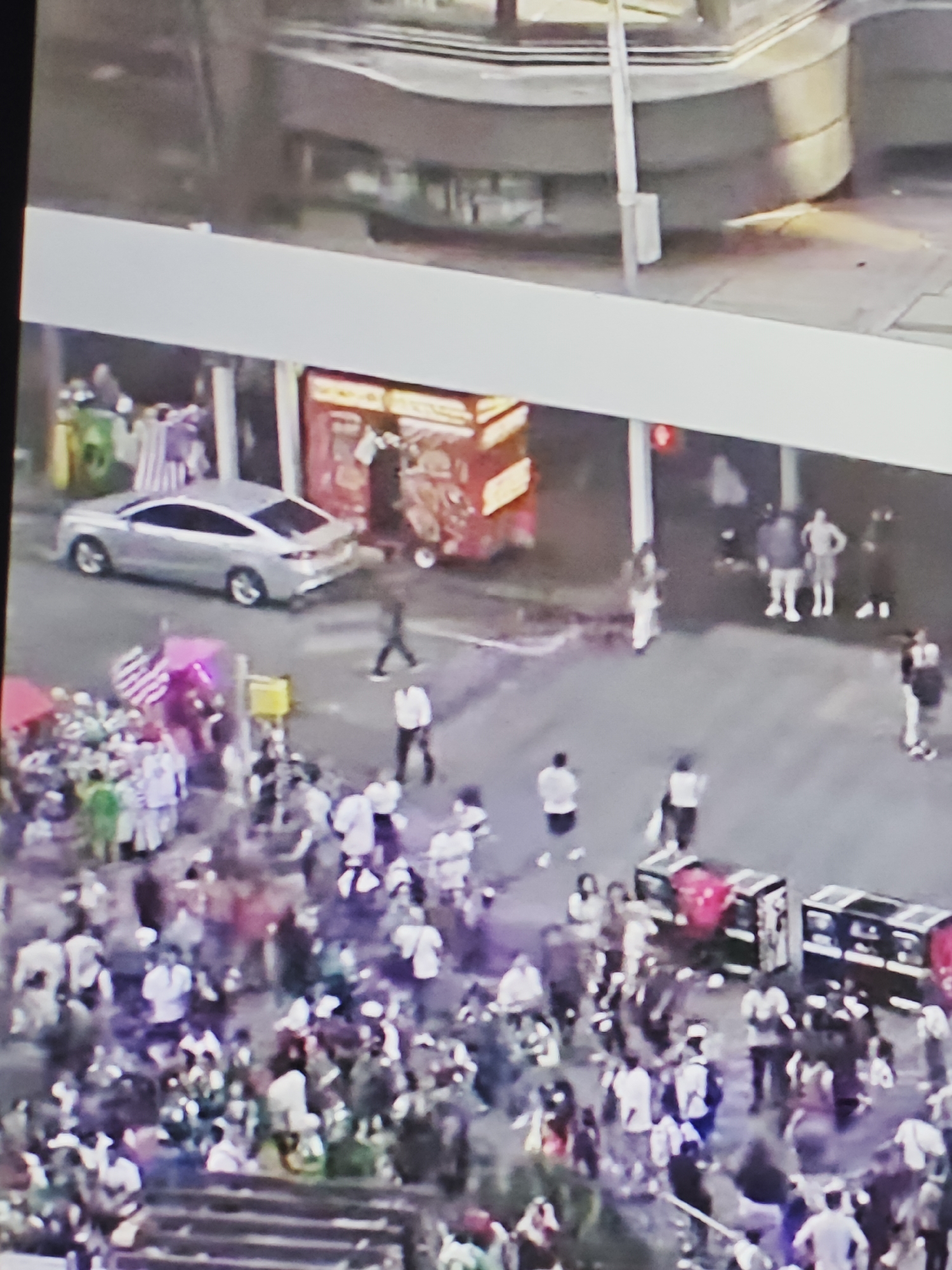 An aerial view captures a bustling outdoor scene, likely in a city. In the foreground, a dense crowd of people is gathered, their forms indistinct due to the distance and low resolution. Many individuals are wearing light-colored clothing. Some appear to be standing or walking amongst the crowd, while others are less discernible.
Behind the crowd, a silver sedan is parked on a street. Further back, a red food cart or stall is visible, with a few people standing nearby. To the left of the food cart, there are some stacked items, one appearing to be a striped bag.
In the background, the scene transitions to what looks like an urban structure, possibly a building or elevated walkway, with some indistinct figures and what might be traffic signals or lights. The overall impression is that of a lively public gathering, possibly a market, festival, or street event. The lighting suggests it is daytime.