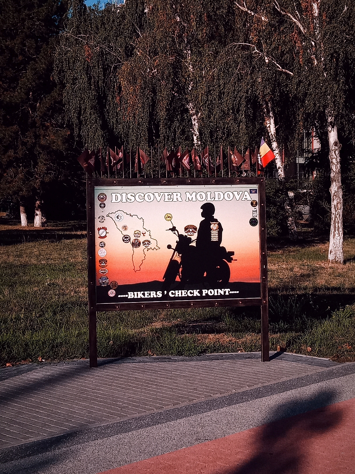 A sign stands in front of a grassy area with trees. The sign has a map of Moldova with several circular emblems on it, as well as a silhouette of a motorcyclist. The text on the sign reads "DISCOVER MOLDOVA ---BIKERS' CHECK POINT---". A Romanian flag is visible to the right of the sign, flying from a flagpole. The ground in front of the sign is paved with rectangular stones. The trees in the background appear to be birch trees.