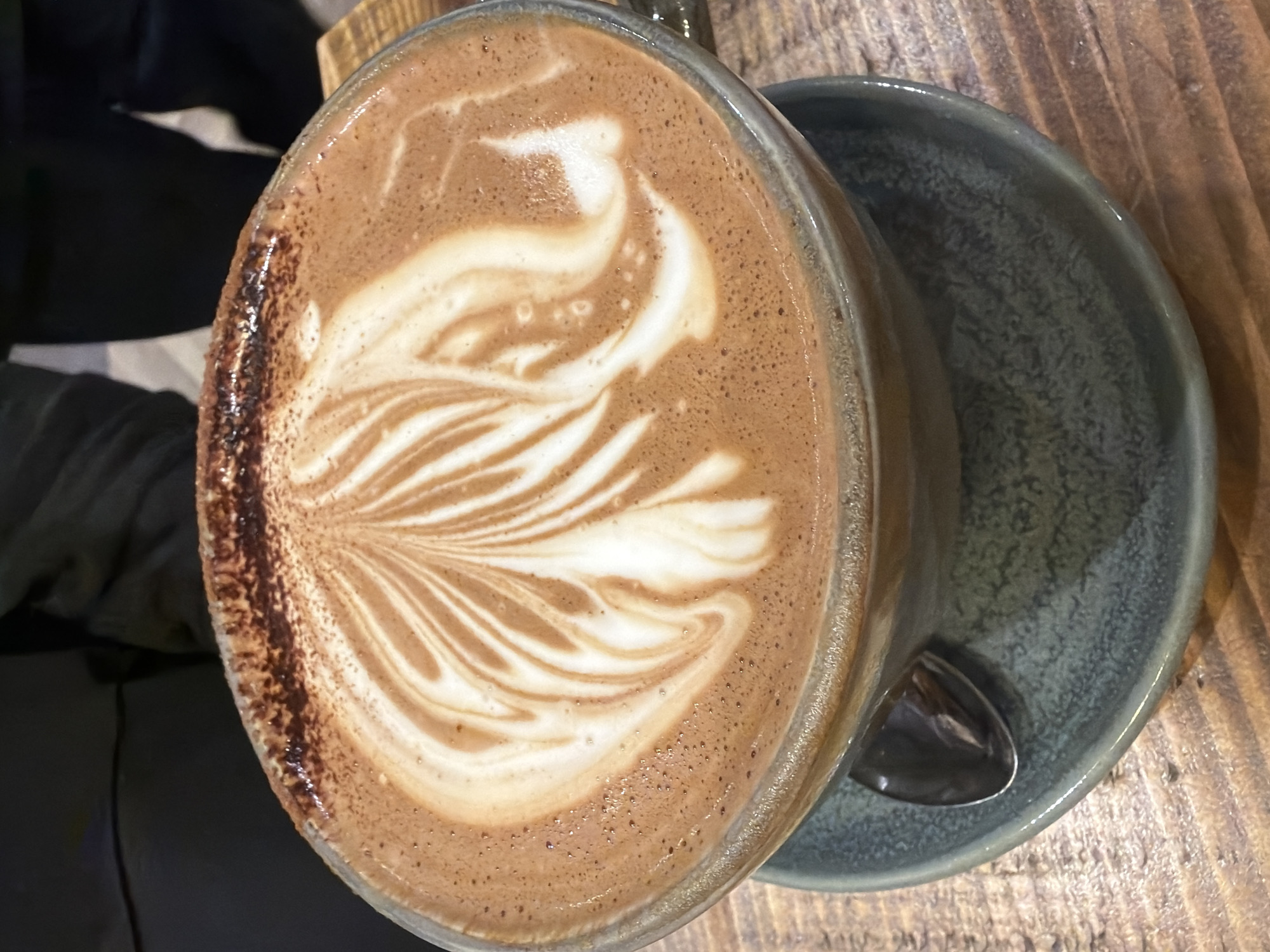 A close-up, angled shot focuses on a cup of latte art resting on a blue-grey saucer placed on a wooden surface. Part of a black-clad individual is visible to the left. The coffee cup is filled with a beverage topped with an intricate white pattern resembling a leaf or feather design. The cup's rim appears dusted with cocoa powder. A small spoon sits in the saucer. The environment suggests a cafe or restaurant setting, possibly in Mole Valley, United Kingdom.
