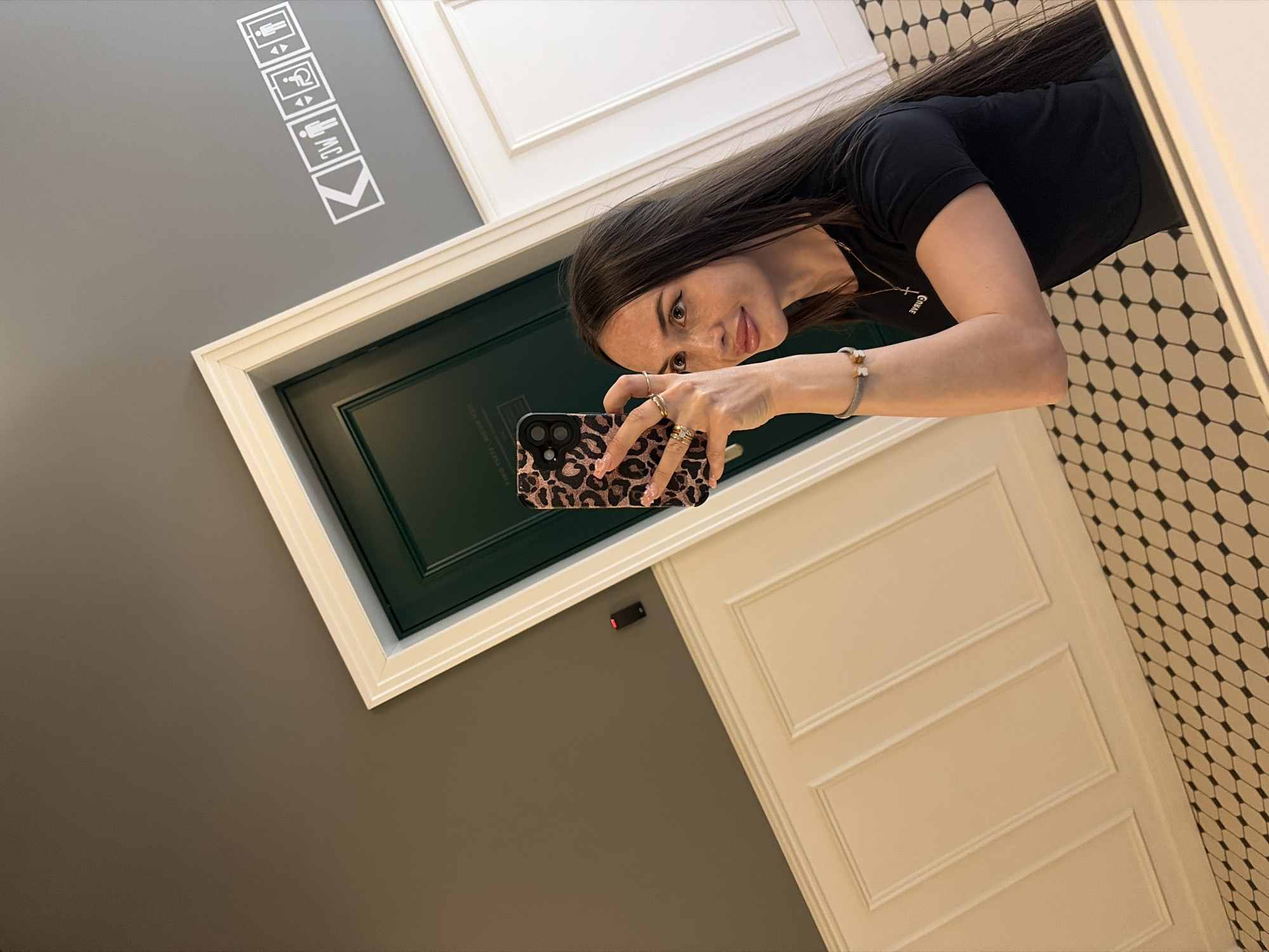 A woman with long, dark hair and fair skin is taking a mirror selfie in what appears to be a public restroom or hallway. She is wearing a black t-shirt and several rings. Her right hand is holding a phone with a leopard-print case, and her left arm, adorned with a bracelet, rests on her hip. The background features a dark green door with a white frame, paneled walls painted cream, and a grey upper wall. On the grey wall, there are white pictograms indicating an elevator and restrooms. A small black electronic device with a red light is mounted on the wall to the left of the door. The floor is tiled with a geometric pattern of black and beige shapes. The mirror's reflection captures the scene from the chest up. The image provides no specific location information for Saint Petersburg, Russia.