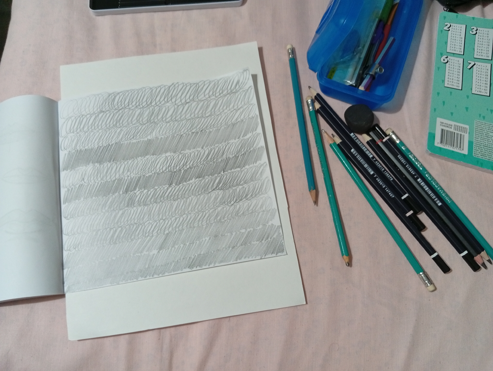 The image shows an open sketchbook with a page filled with pencil strokes that create a textured, shaded effect. Several pencils and an eraser are scattered around the sketchbook on a pink fabric surface. A blue pencil case, filled with more pencils and writing utensils, is also visible. In the background, a green paper with multiplication tables is partially visible. The scene appears to be a tabletop or desk in a home environment. There is no discernible information in the image that would allow for the localization of this scene to San Antonio de Los Altos, Venezuela.