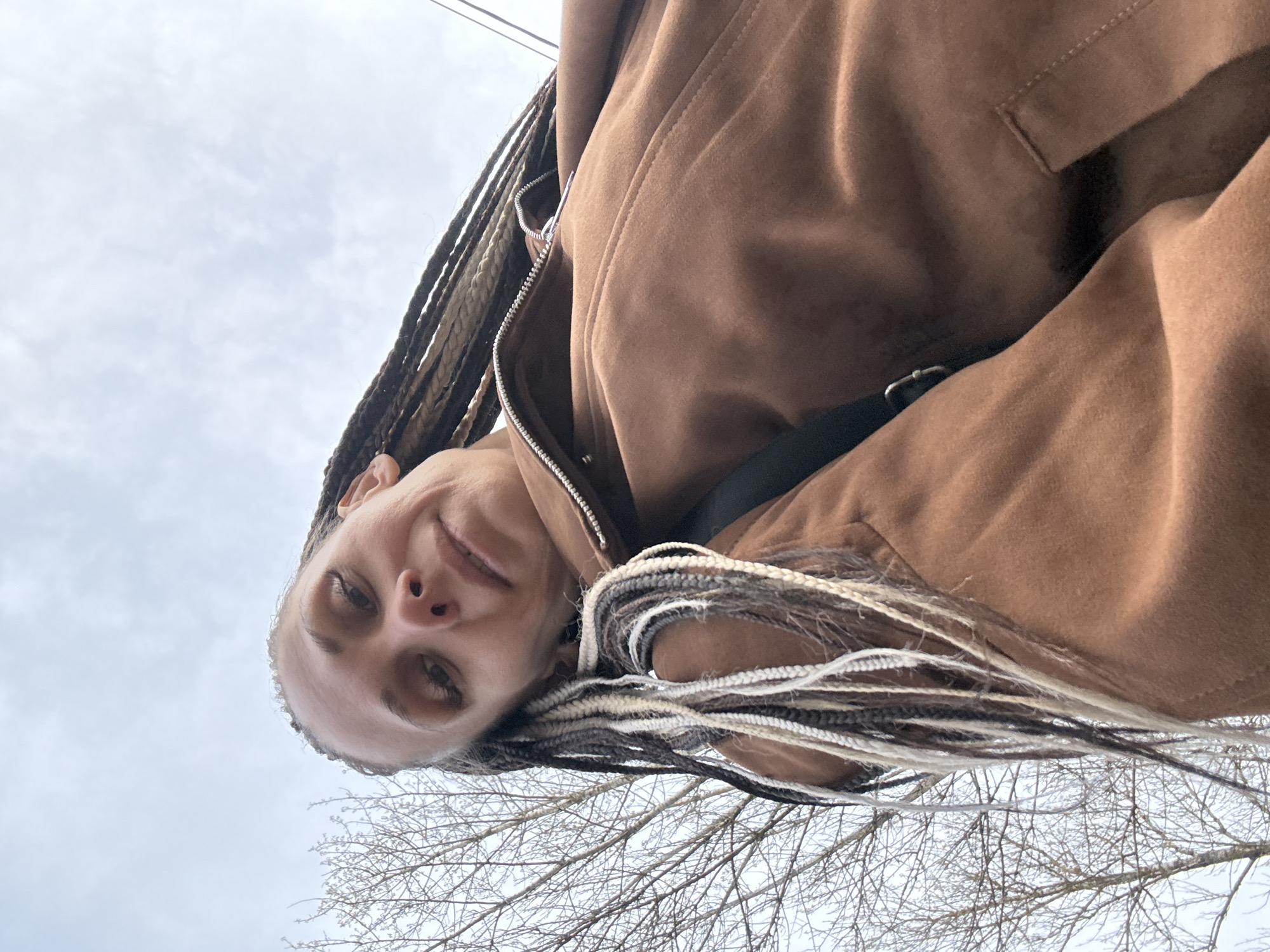 A low-angle, tilted selfie photograph depicts a woman with braided hair (primarily dark, with some white strands interspersed) wearing a brown jacket. The jacket appears to be partially zipped. A thin black strap is visible beneath the jacket, likely from a bag or purse. The background consists of a cloudy sky with bare tree branches in the lower right corner. The location is Rusanovo, Russia.
