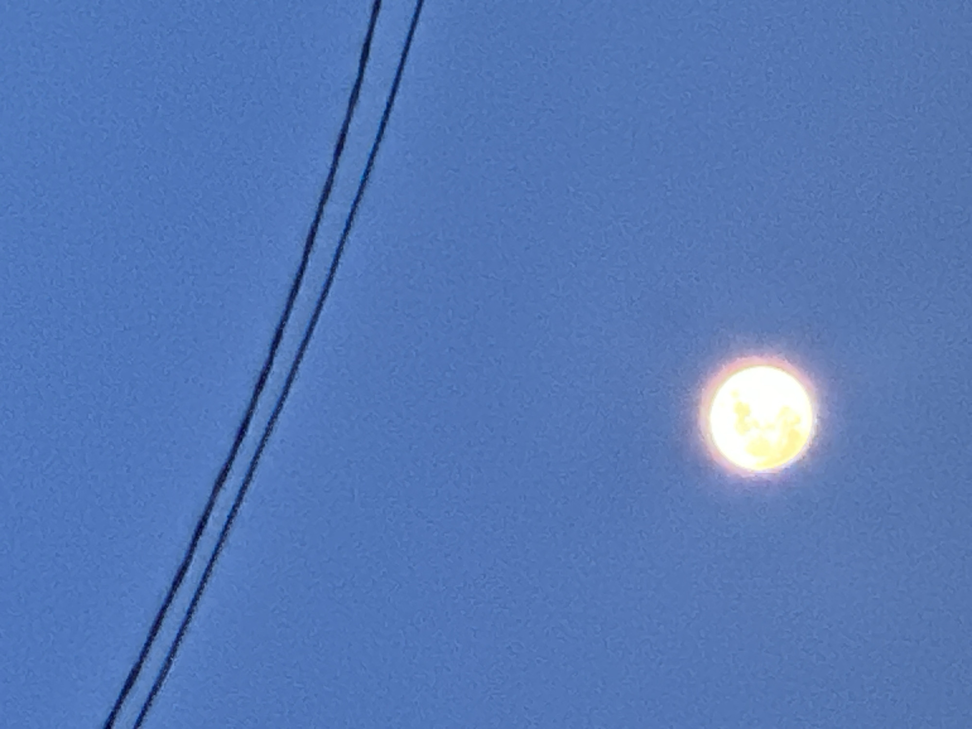 The image depicts a full moon in a blue sky, with electrical wires visible in the upper left corner. The scene is likely set in Chaikovsky, Russia, but there are no visual cues in the image to confirm this information. The focus is on the moon, illuminated against the blue sky. No people or objects are visible other than the moon and the wires.