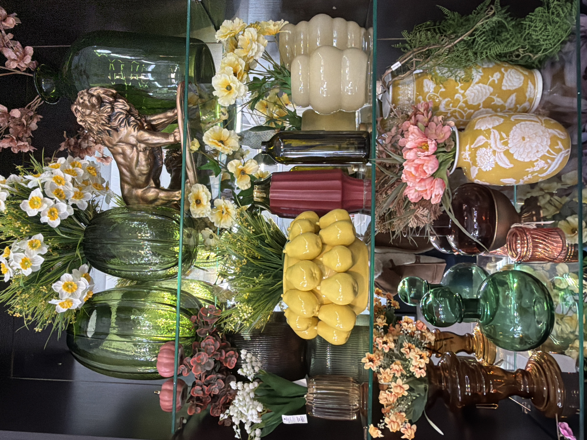 This image captures a multi-tiered retail display shelf, likely within a home decor or gift shop in Votkinsk, Russia. The background is uniformly dark, causing the illuminated objects on the clear glass shelves to stand out.
The **top shelf** features two large, translucent dark green ribbed glass vases flanking a bouquet of artificial white and yellow daffodils with green foliage, presented in a similar green ribbed vase. To the right of the daffodils, a bronze-colored statue of a standing lion faces right. A tall, dark green glass vase stands to the lion's right. Two small, matte pink pumpkin-shaped decorative objects are positioned in the foreground on the left, and a cluster of dried or artificial reddish-brown floral branches is visible in the upper right.
The **middle shelf** displays a clear, ribbed glass vase containing artificial white lilies-of-the-valley and green leaves on the far left. Behind it, a tall, dark brown ribbed vase is partially visible. Centrally located is a bright yellow ceramic vase, uniquely textured with multiple pear or lemon shapes, filled with green foliage and small yellow flowers. To its right, a tall, ribbed, dark red ceramic vase stands beside a slender, dark brown glass bottle. Further right, two off-white, multi-sectioned (segmented) ceramic vases hold artificial yellow and white flowers. Reflections on the shelf's surface indicate the presence of a person, identifiable by a brown jacket.
The **bottom shelf** on the left holds a dark brown ornate vase containing artificial peach-colored flowers and green foliage, accompanied by a smaller, similar vase. Adjacent to these are two dark green glass decanters with stoppers; the left one is spherical, and the right is an elongated oval. A small, ribbed, rose-gold colored glass votive or jar is placed to their right. Next are two tall, yellow ceramic vases with embossed white floral patterns, containing artificial pink tulips. To the far right, a trailing arrangement of artificial green ferns cascades downwards. Reflections of the surrounding retail environment, including partial figures, are visible across the glass surface.