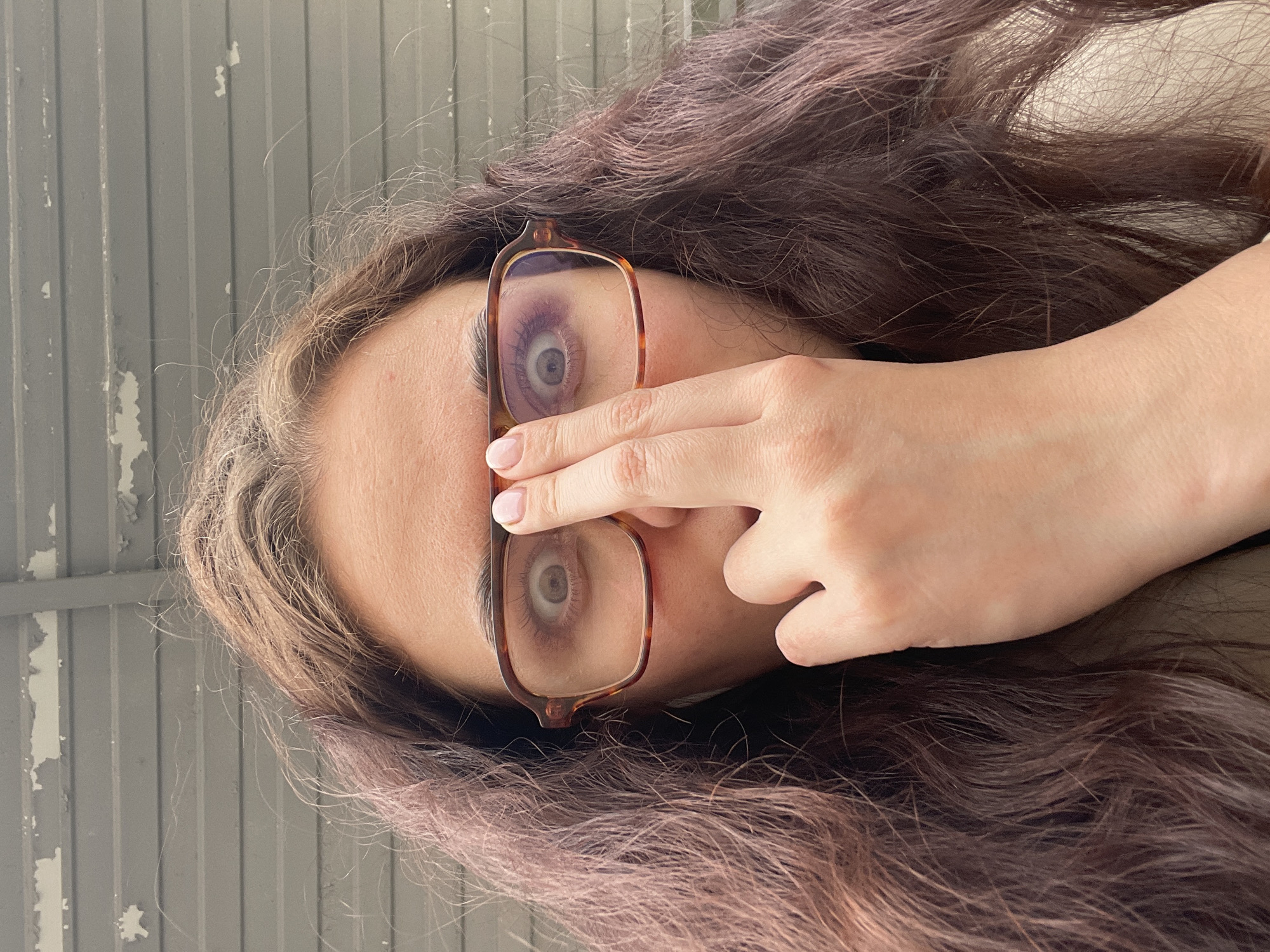A woman's face is seen through eyeglasses, with her hand partially obscuring her forehead. She has long, wavy, brown hair. The background appears to be a painted wall with flaking paint. The location is unknown.