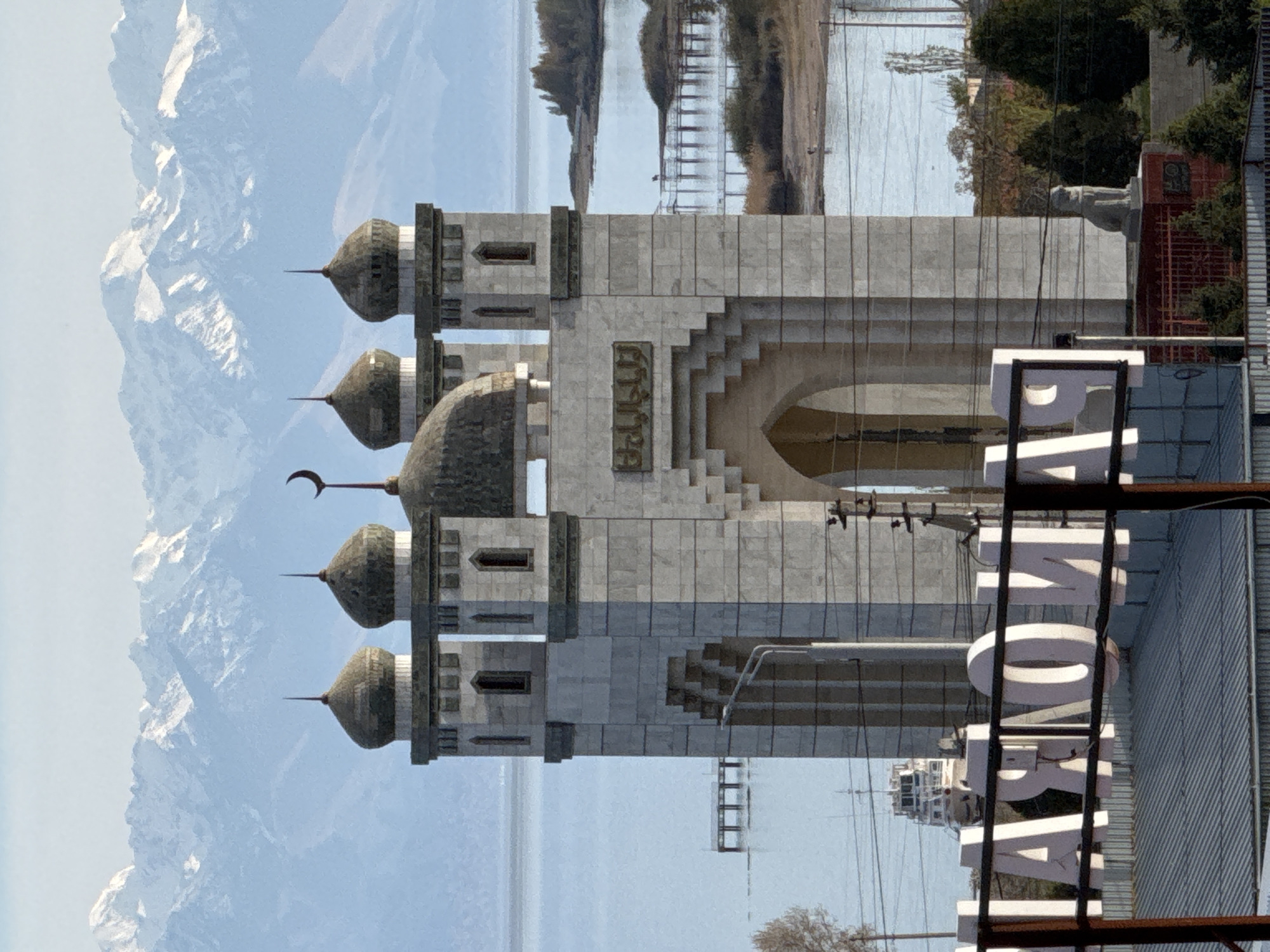 The image shows the Rukh Ordo Cultural Center in Çolpon-Ata, Kyrgyzstan. In the foreground is a partial view of the "PAMOMAPA" sign. The main subject is the building of the cultural center, featuring multiple domes and arched structures. In the background, there are snow-capped mountains and what appears to be a body of water. Trees and some infrastructure are visible as well.