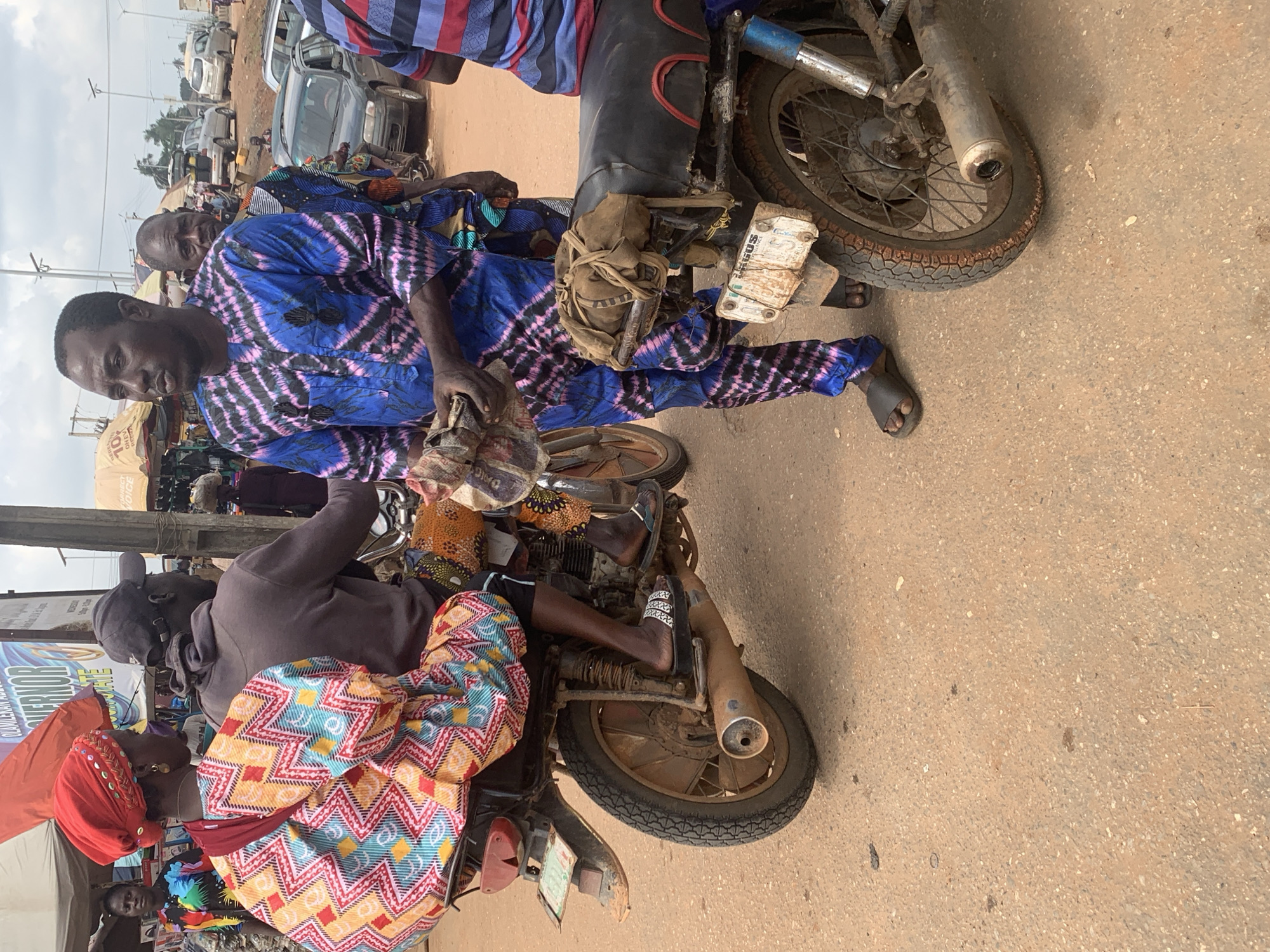 In Shagamu, Nigeria, a man in a vibrant blue and pink tie-dye dashiki stands on a dirt road beside a motorcycle. He is holding a bundle of what appears to be wrapped goods in his hands. Behind him, a woman wearing a red headscarf and a colorful patterned dress is seated on the back of the same motorcycle, her legs tucked to the side. The motorcycle is worn and dusty, with a visible exhaust pipe and a license plate that reads "LAGOS". In the background, other people, motorcycles, and cars are visible, suggesting a busy marketplace or street scene. The sky is overcast, and the overall environment appears to be outdoors in a bustling urban area.