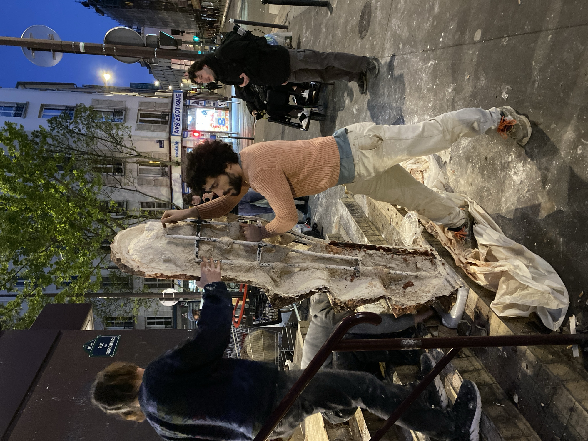 The image shows a group of people working on a large, irregularly shaped object in an urban environment, specifically Paris, France, as indicated by the street sign "Rue des Poissonniers". One person is prominently featured, leaning over the object, wearing a peach-colored sweater and light-colored pants, which are splattered with white material, suggesting artistic activity. Another person is positioned at the bottom of the image, facing away from the viewer, wearing a dark jacket. In the background, a third individual is visible, possibly operating a camera or other equipment. The scene is set against a backdrop of Parisian buildings and street signage. The irregularly shaped object appears to be a sculpture or a mold, covered in a white, plaster-like substance and reinforced with metal. The ground around the object and the individuals involved is also marked with splatters of the same white substance. Overall, the image captures a scene of artistic creation or restoration taking place in the public space of Paris.