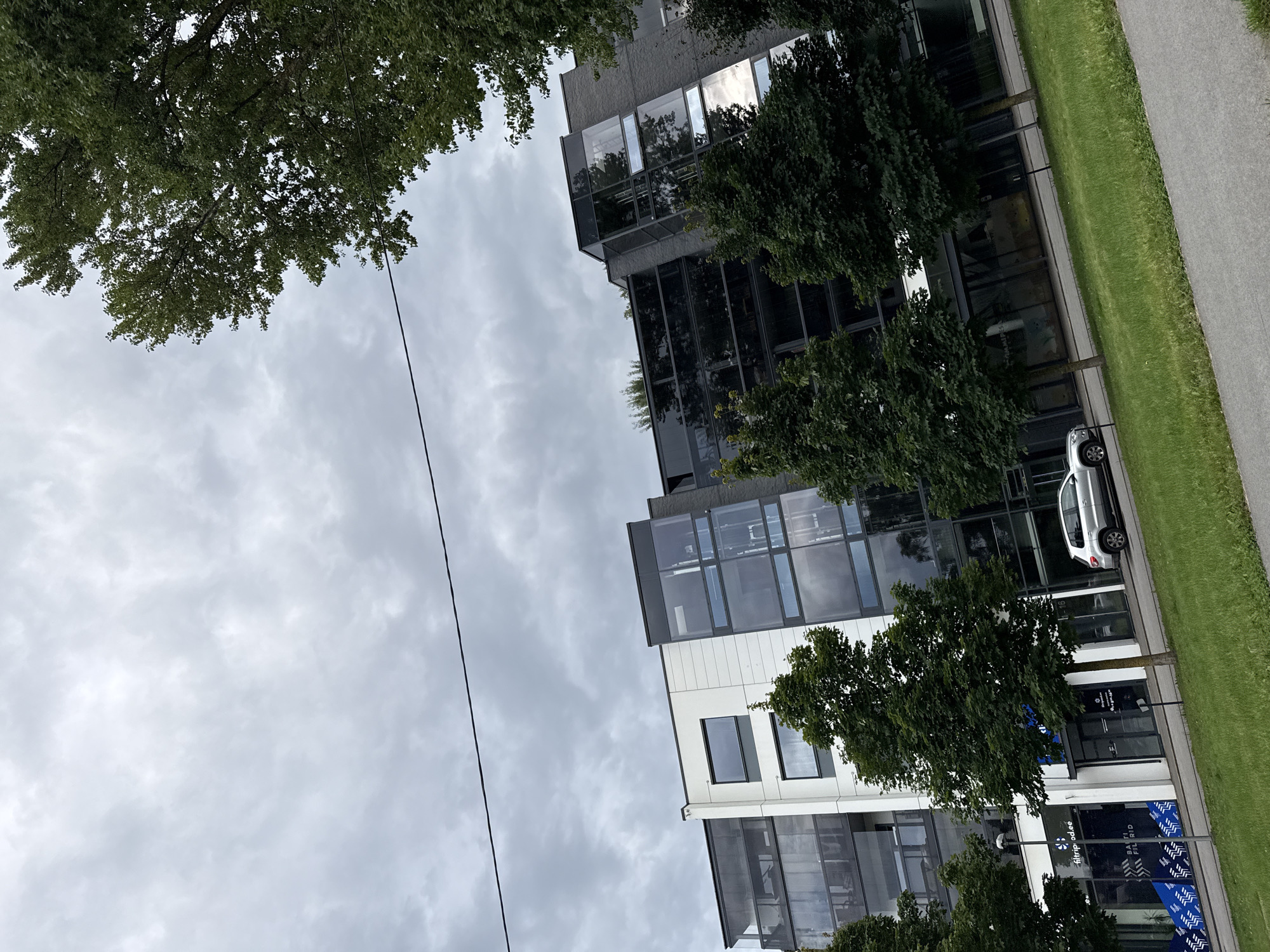 The image depicts a modern apartment building in Tallinn, Estonia, with a silver SUV parked in front. The building features a contemporary design with white and grey facades, large glass windows, and balconies. Lush green trees line the street in front of the building, and a grey asphalt path is visible in the foreground. The sky is overcast, suggesting a cloudy day. The building is located on a street in Tallinn, Estonia.