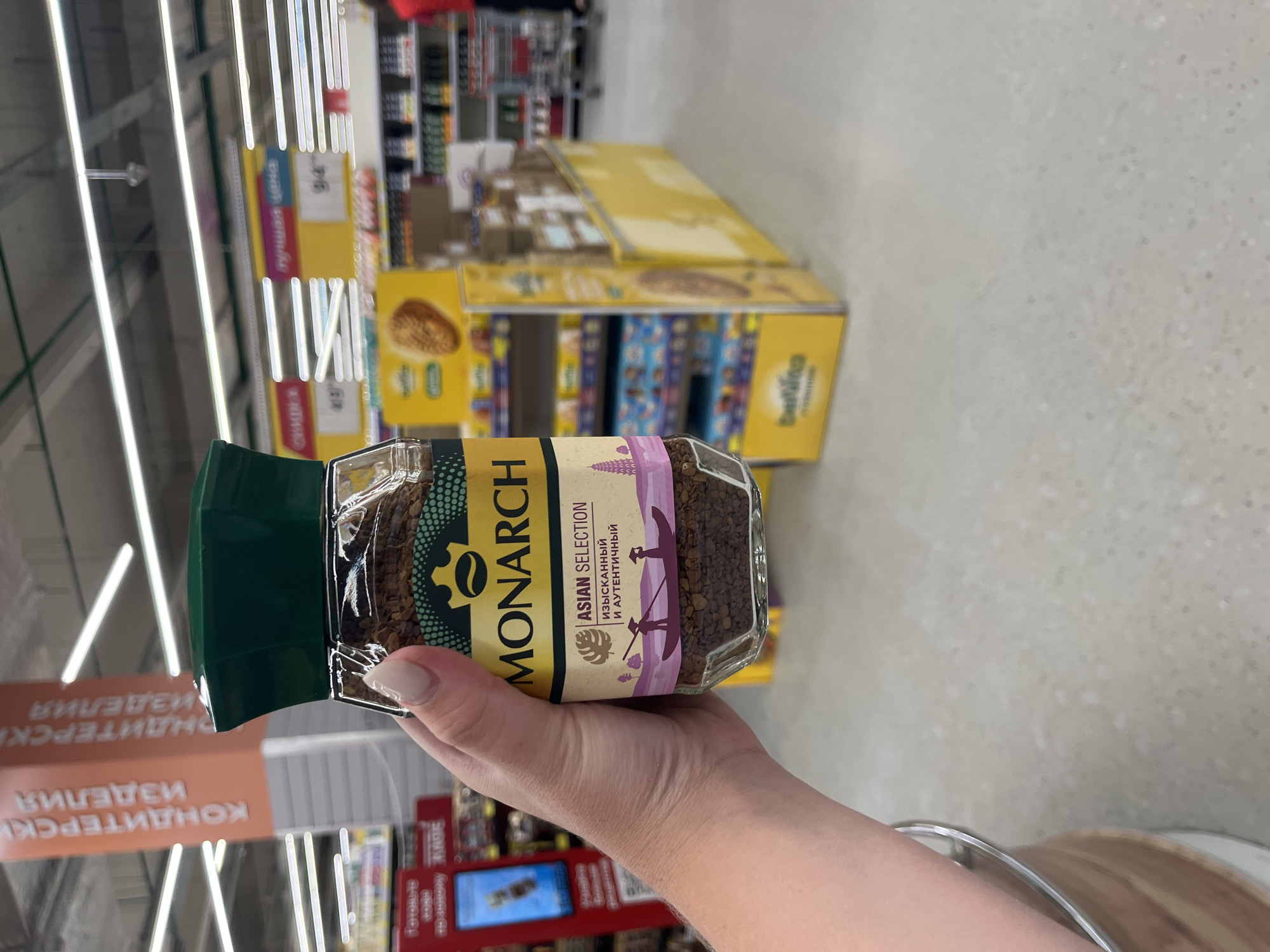 A person's hand, with painted fingernails, holds a glass jar of Monarch brand instant coffee. The jar is labeled in English and Russian. The background shows a retail environment, likely a supermarket in Volzhsky, Russia, with aisles of products, promotional displays, and overhead lighting. A sign with Cyrillic text, possibly indicating "Confectionery Products," is visible in the upper left. A price tag with "94" is displayed on a yellow promotional stand. The floor is a light-colored speckled material.