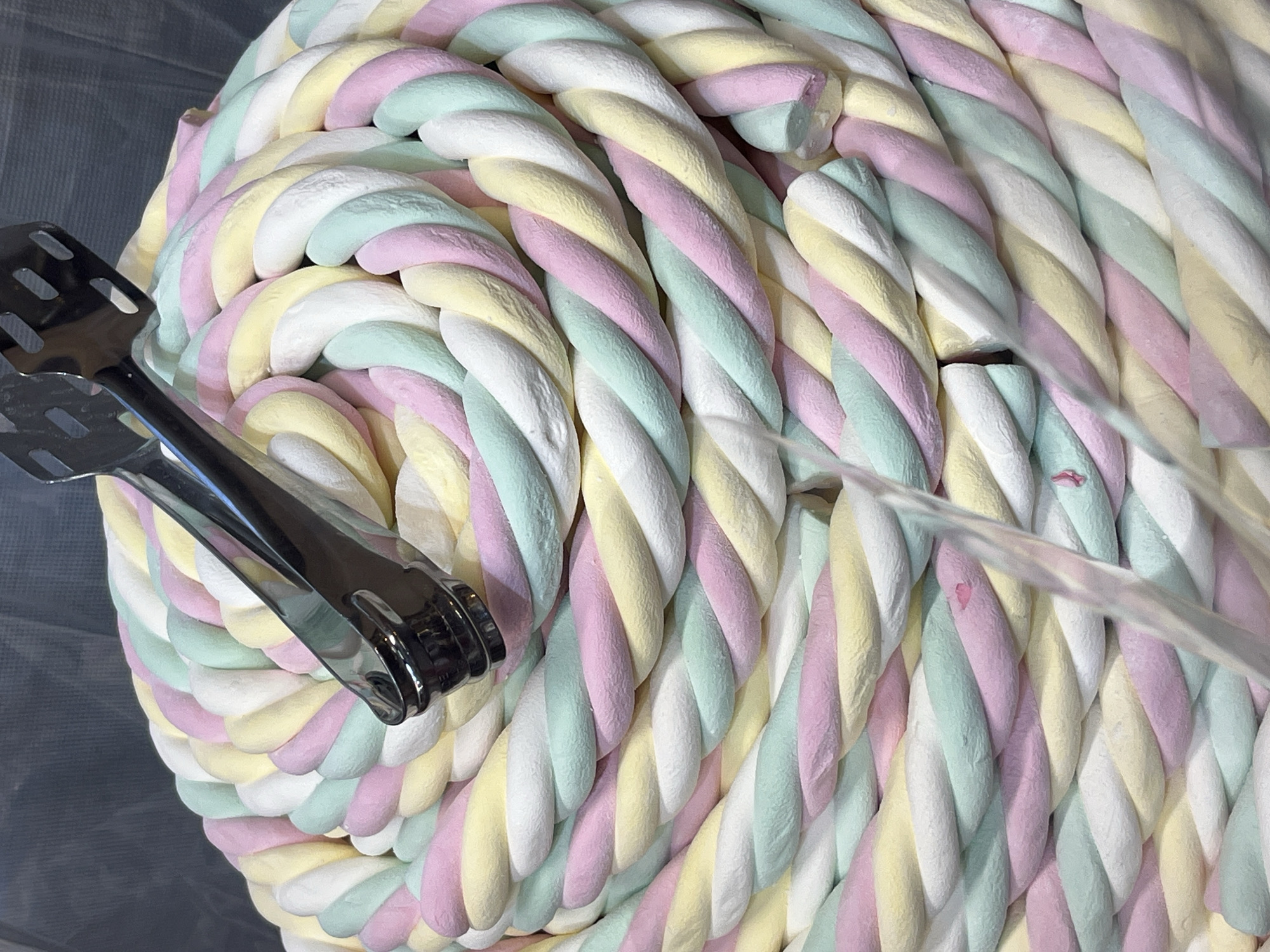 A close-up, high-angle shot depicts a large pile of twisted marshmallows in pastel colors of white, yellow, pink, and mint green. A shiny, metal serving tong is placed to the left of the frame, resting atop the marshmallows. A blurred, transparent object, possibly a plastic divider, cuts across the right side of the image. The pile is set in a tray of some kind, set to the right of the frame. The marshmallows appear to be coiled tightly together. The specific location in Budva, Montenegro, cannot be determined from the image. No people or interactions are visible.