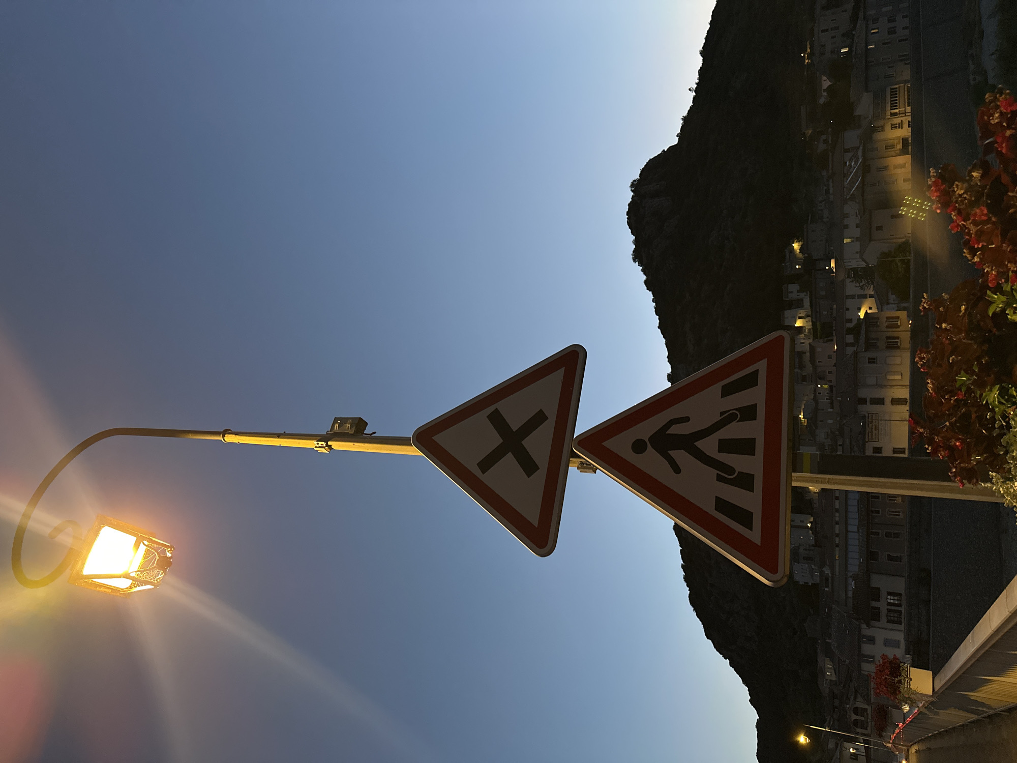 The image depicts a street scene in Anduze, France, at dusk. Two triangular road signs are prominent: the upper one displays a black "X" indicating an intersection, while the lower one shows a silhouette of a pedestrian crossing a crosswalk. Both signs have red and white borders. They are mounted on a yellow pole, which also supports a street lamp emitting a warm, yellow light. The sky is a clear, deep blue, transitioning from lighter shades near the horizon. In the background, a dark, tree-covered mountain looms over a village. The village's buildings are visible, with some lights illuminated in windows. The foreground features a railing, possibly part of a bridge or walkway, with planters containing red flowers. The overall atmosphere is serene, marking the transition from day to night.