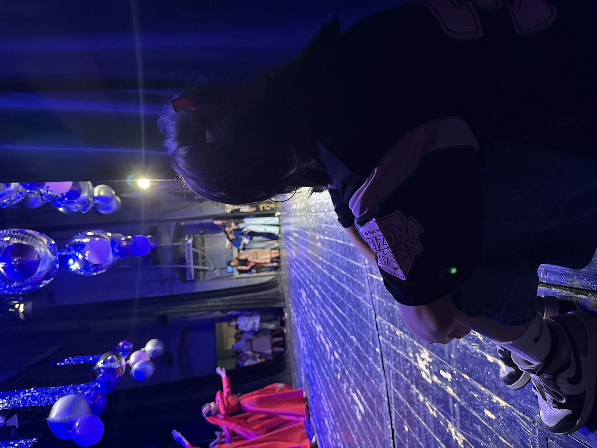 A vertical shot, likely taken by a person standing on a stage, shows a performance being viewed. The photographer is wearing sneakers, blue jean shorts, and a black top with white accents and a patch that says "Offensive Line Arizona Stars 1992." They are looking down at the performance, which involves several people dressed in red costumes. Blue and white balloons decorate the stage area. In the background, there is a second-story balcony where more people are standing and watching the performance. The scene appears to be indoors and brightly lit with blue stage lights. The location of the city of Yerevan, Armenia cannot be confirmed using only the provided image.