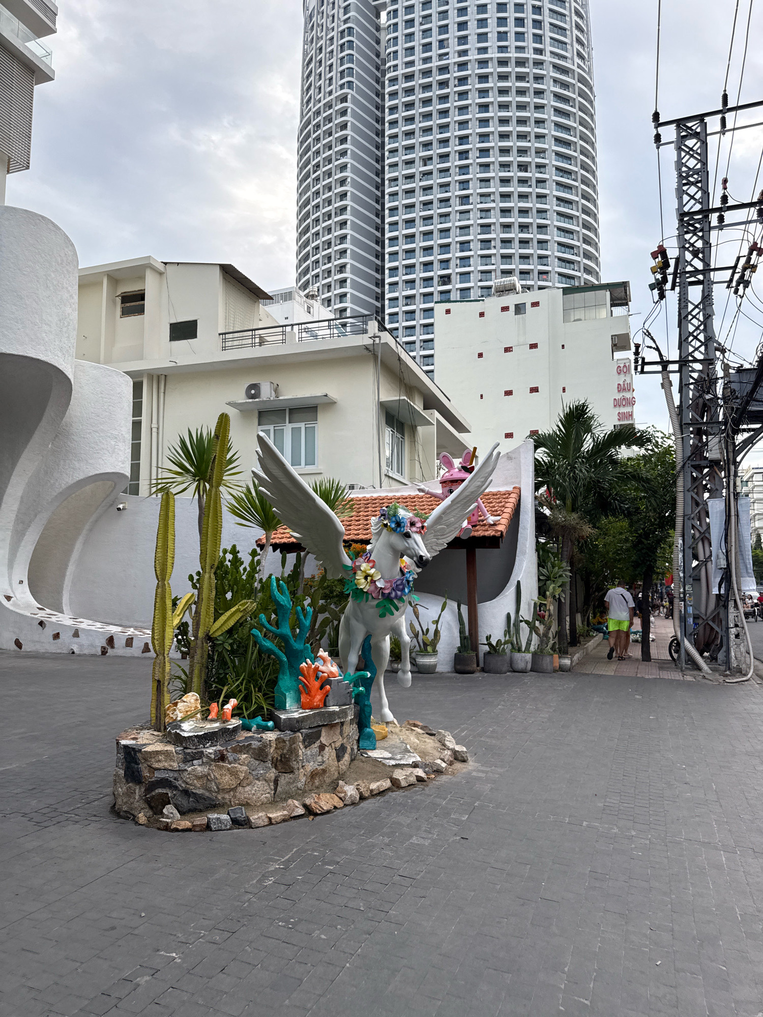 In front of a tall, modern skyscraper in Nha Trang, Vietnam, a white statue of a winged horse adorned with a flower wreath is positioned in a landscaped area. The statue stands on a stone base decorated with colorful coral figures. Behind the statue, a white building with a tiled roof is visible, and further back, a multistory skyscraper dominates the skyline. On the right side of the image, a utility pole with numerous wires stands prominently. Two individuals are walking on a paved walkway towards the right of the frame, and a motorcycle is parked near the pole. The ground is paved with dark gray interlocking stones, and several potted plants, including cacti, are scattered throughout the scene. The sky is overcast, suggesting a cloudy day.