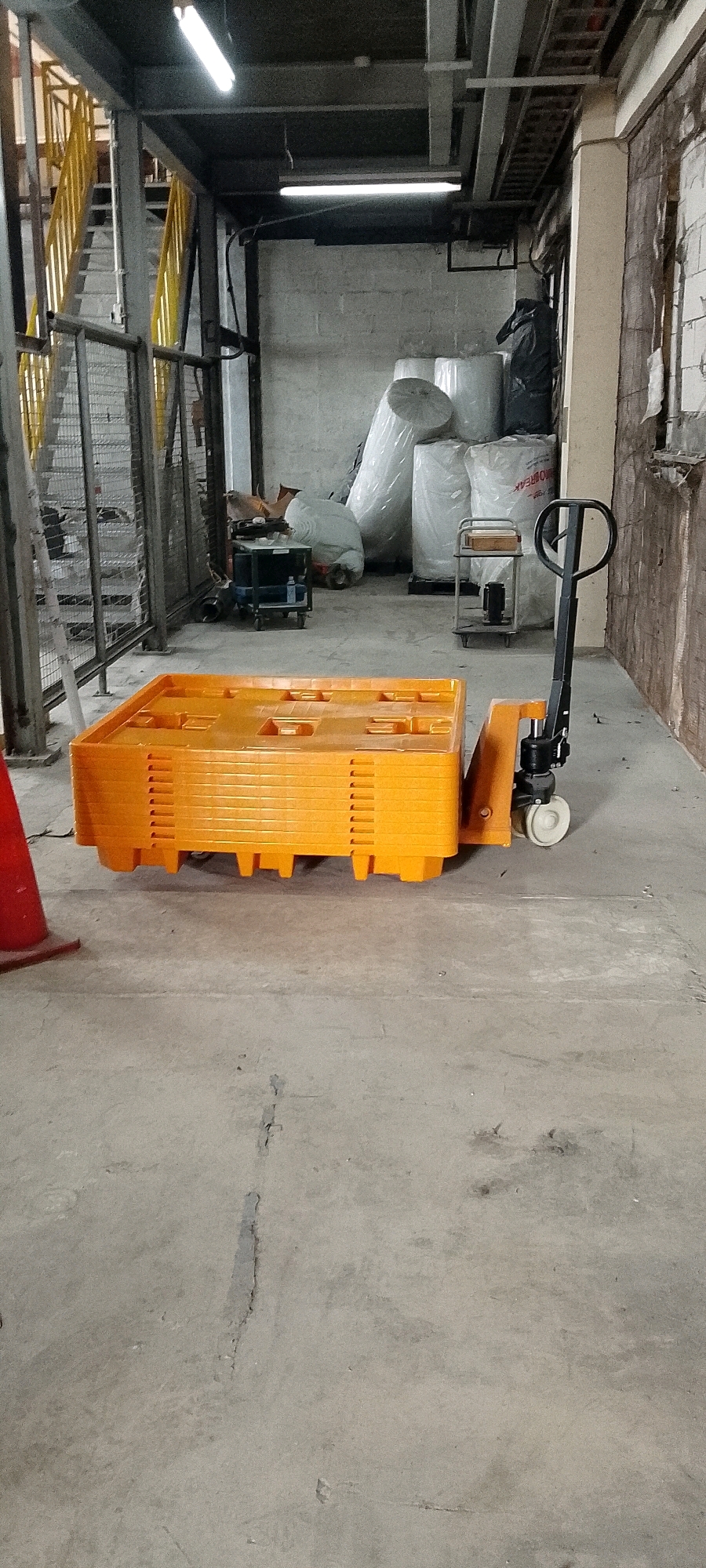 In Depok, Indonesia, an indoor scene depicts a concrete floor in what appears to be a storage or industrial area. In the foreground, a stack of bright orange plastic pallets rests on a pallet jack. The pallet jack has a black handle and white wheels. To the left of the pallets, a portion of a red traffic cone is visible. In the background, there are various objects including large white rolls of material wrapped in plastic, a black trash bag, and a metal cart with shelves. A metal staircase with yellow railings is visible on the left side of the image. The walls are made of concrete blocks, and fluorescent lights are installed on the ceiling.