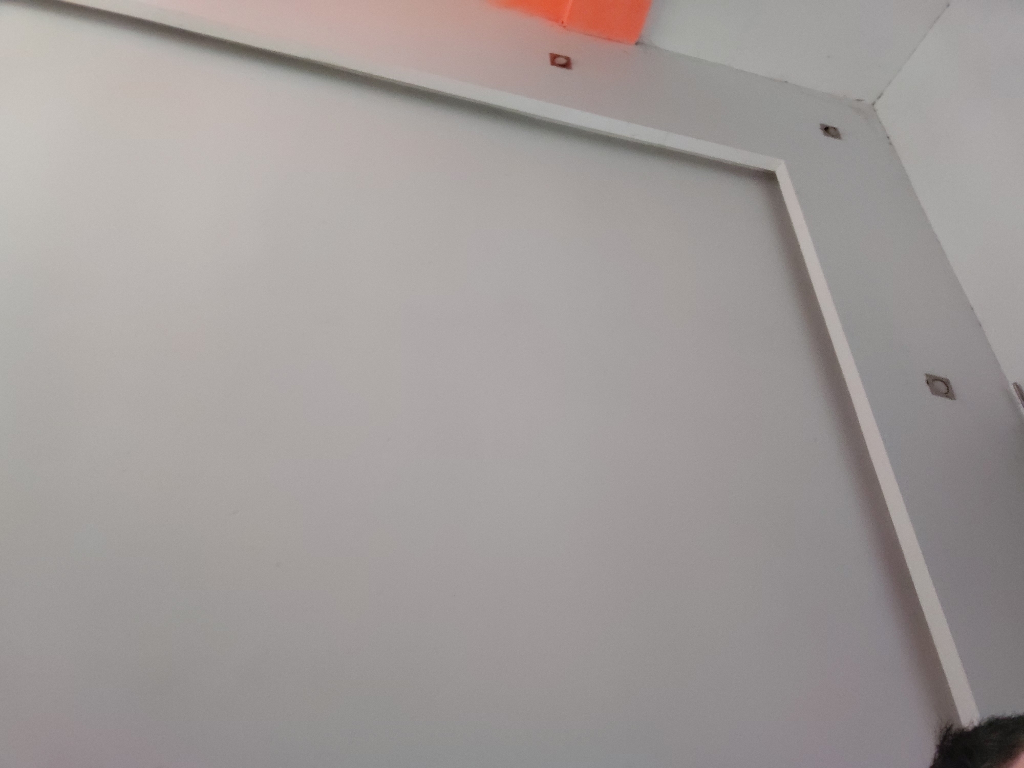 The image displays an interior scene, likely a room or hallway in Santa Ana, Venezuela. The central subject is a large, light grey rectangular panel or wall section, bordered by white trim along its top and right edges. Above the top trim, an orange object or material is visible against the ceiling. Several small, dark, recessed holes—some square, others circular—are present on the wall and ceiling near the upper right corner, possibly for electrical outlets, lighting fixtures, or mounting purposes. The corner where the two walls and ceiling meet appears somewhat irregular or unfinished. In the bottom right corner of the frame, a small section of dark hair, presumably belonging to the person taking the photograph, is visible. No explicit activity is taking place, but the appearance of the fixtures and wall suggests the space might be under construction, renovation, or a functional area. The lighting is even, typical of an indoor setting. There is no visible text.