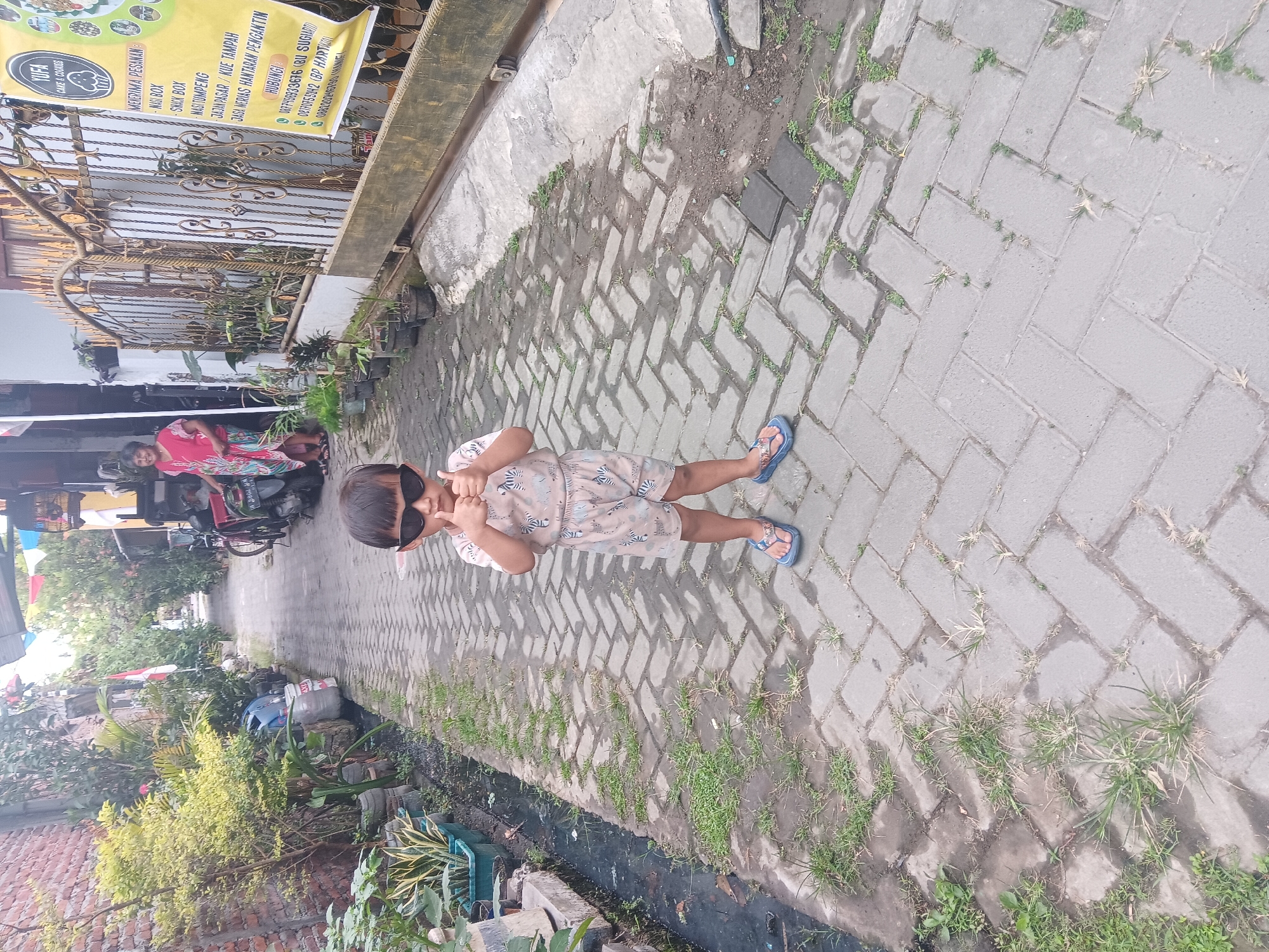 A young child wearing sunglasses and a patterned outfit stands on a paved walkway in the city of Tlogo, Indonesia. The child is giving a thumbs-up with both hands. To the right of the child, a metal fence encloses a property with potted plants. A banner is attached to the fence, featuring text in Indonesian and the logo "YUFA CAKE & COOKIES". In the background, a woman in a pink and floral dress stands near a motorcycle. The walkway is made of gray brick pavers, with patches of grass growing between them. The environment appears to be an outdoor alleyway or residential street, with brick walls and dense greenery visible.