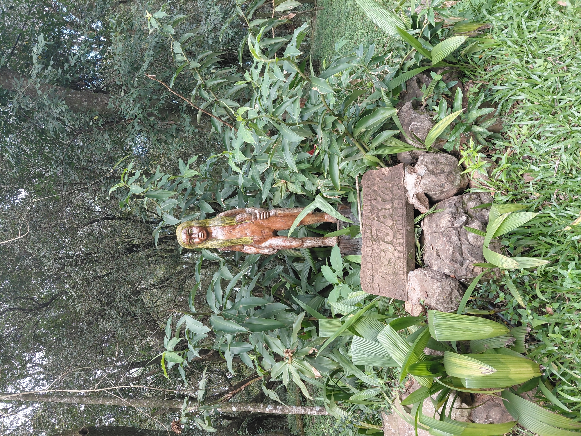 The image shows a wooden statue of a child with long blonde hair standing in a garden. The statue is positioned in front of a lush green foliage of plants and trees, with rocks and grass surrounding its base. A stone plaque with carved inscriptions is visible at the foot of the statue. The inscription on the plaque appears to say "Paseo de los Mitos" and other illegible text. The setting suggests an outdoor location, likely a park or a garden, possibly an exhibit of myths given the inscription. The statue is carved from wood and has a weathered appearance. It depicts a standing figure with its left hand holding what appears to be a small object, and its right hand resting by its side. The statue's face has carved features, with eyes, nose, and mouth visible. The overall scene is one of natural beauty and artistic display, potentially in a public space within Trinidad, Paraguay, as suggested by the plaque's wording possibly referring to "Mitos" (Myths) and a location possibly related to Trinidad.