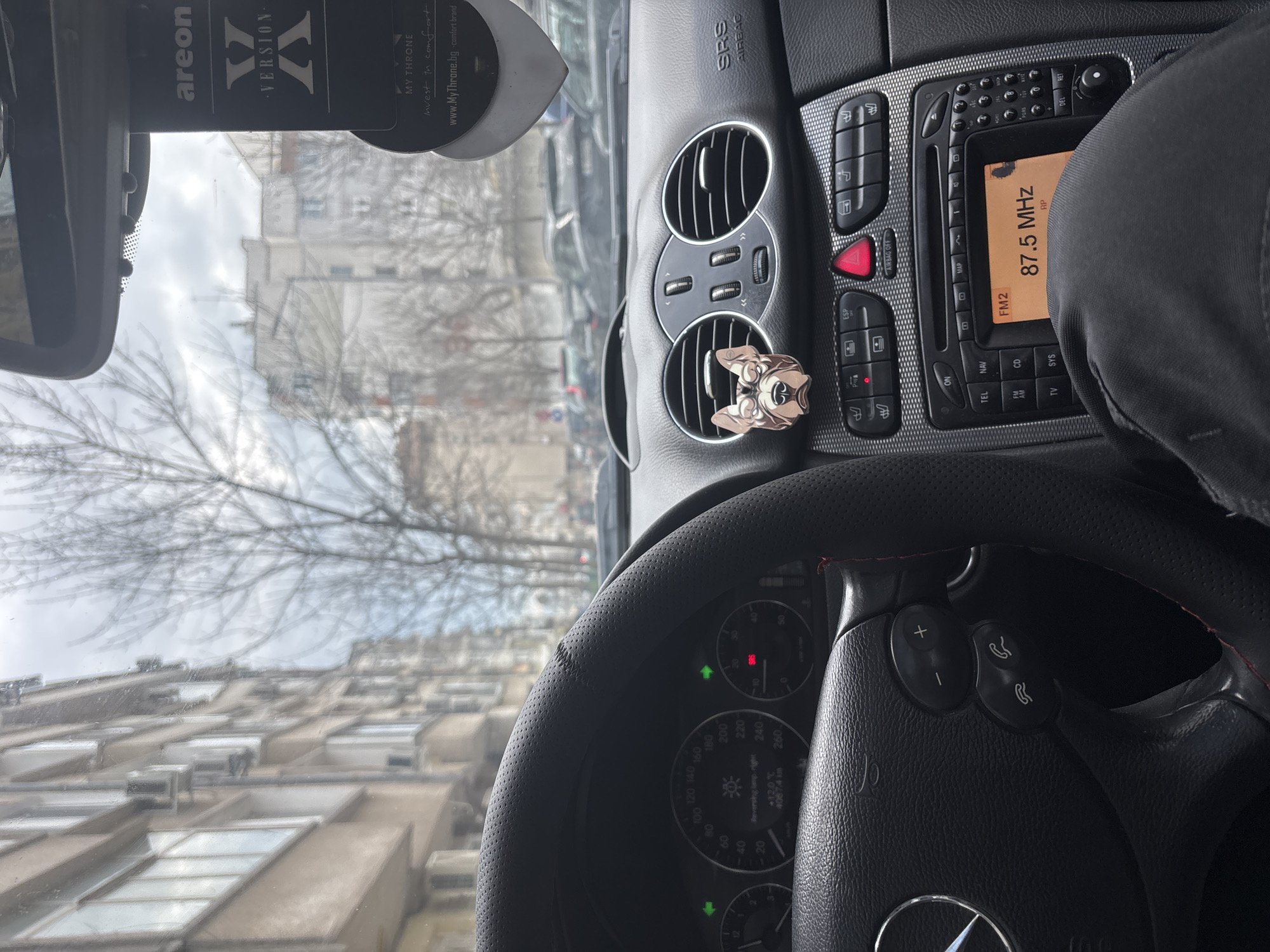 The image shows the interior of a Mercedes-Benz car, likely parked in a city. The steering wheel, dashboard, and center console are visible. The radio is tuned to 87.5 MHz. An air freshener hangs from the rearview mirror. An ornament is clipped onto one of the dashboard air vents. Through the windshield, a building with multiple windows can be seen, suggesting a residential or commercial area.
Based on the air freshener brand "My Throne" with the domain "www.MyThrone.bg" and the fact that the radio is playing an FM frequency, the location is likely in Bulgaria. Burgas is not identifiable as the location based on the information provided in the image.
